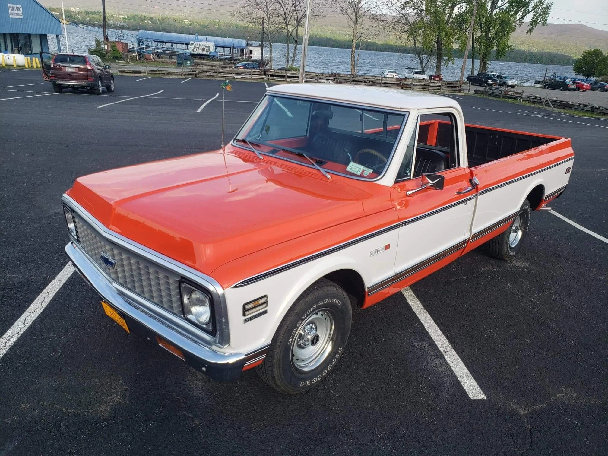 A vintage red and white pickup truck parked diagonally in a parking lot near a body of water, with a blue building and trees in the background.