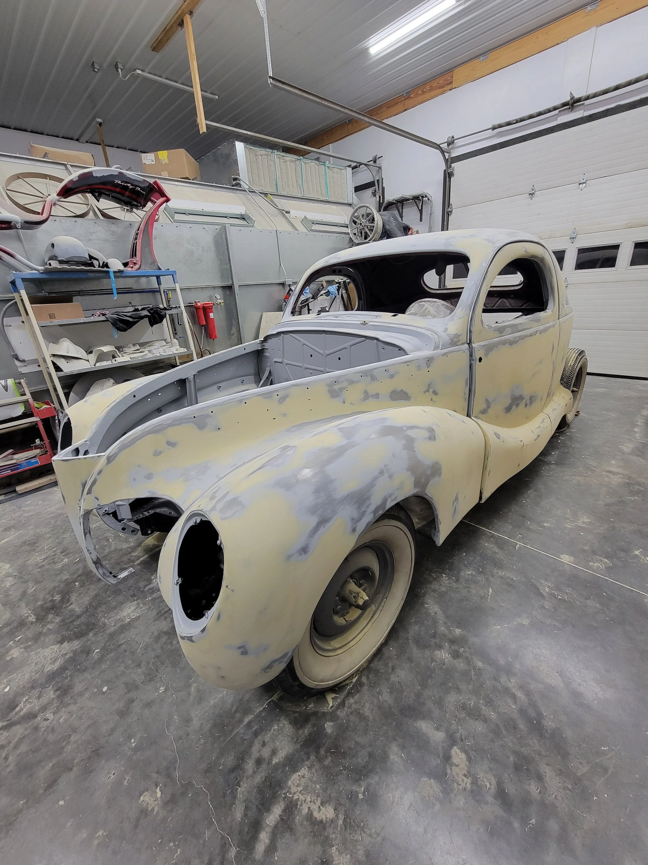 An old vintage car under restoration inside a garage, with a partially painted body and no front lights or grille.