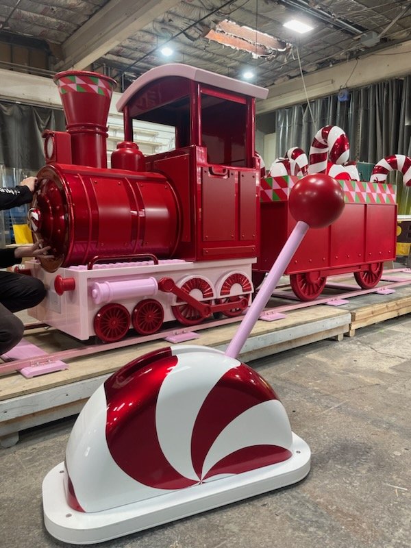 A large red and pink holiday-themed train decoration with candy canes in the background and a large peppermint candy-shaped lever in the foreground.
