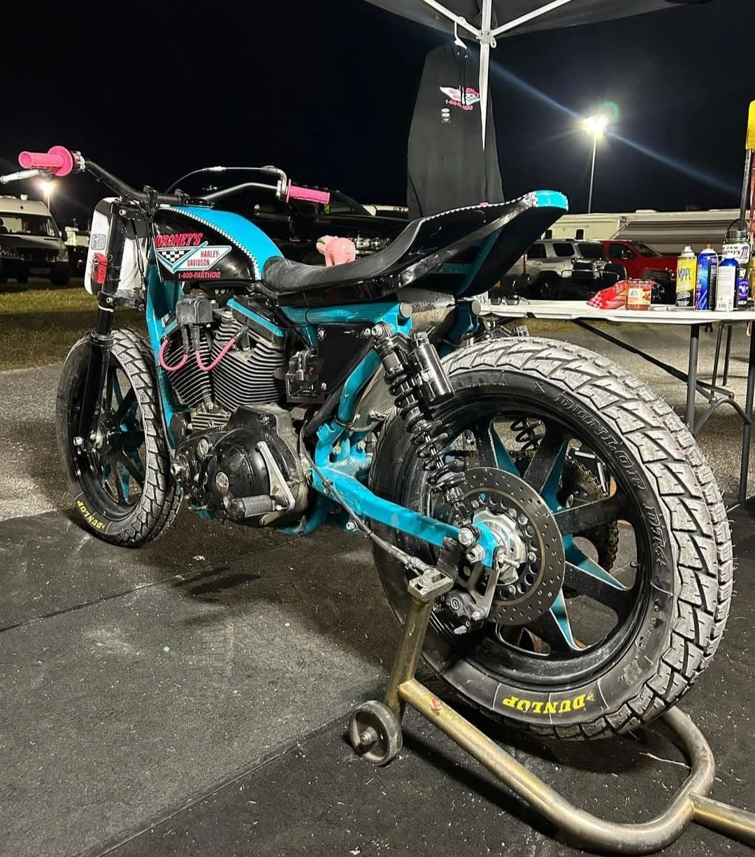 A motorcycle on a paddock stand at night, with a black seat, teal frame, racing tires, and visible engine and suspension components. In the background, a table with products and a black shirt hanging, with cars and bright lights in a parking lot sett