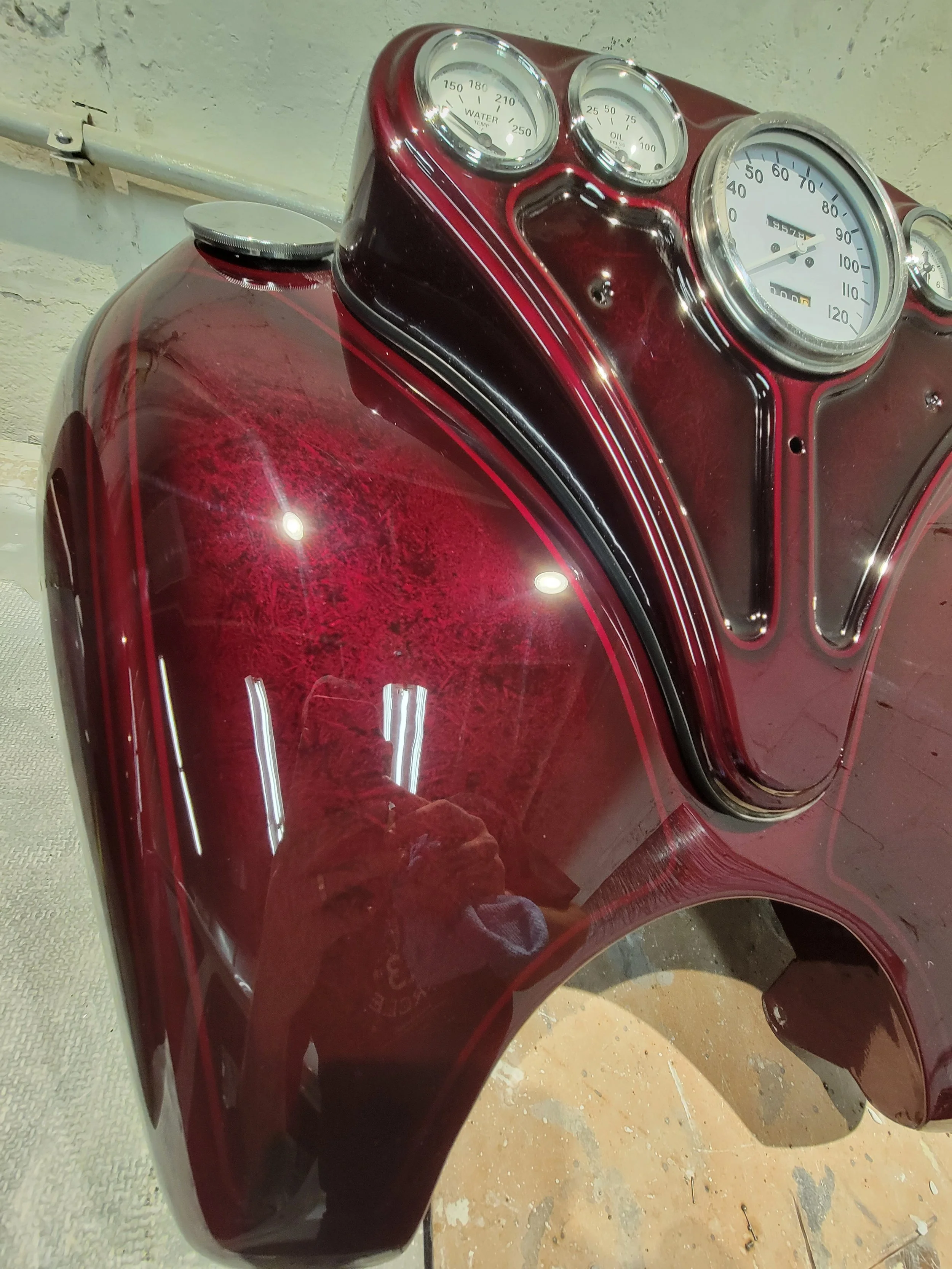Close-up of a vintage motorcycle's red gas tank and dashboard showing gauges for water temperature, oil pressure, and speed. The reflection of the photographer is visible on the shiny surface.