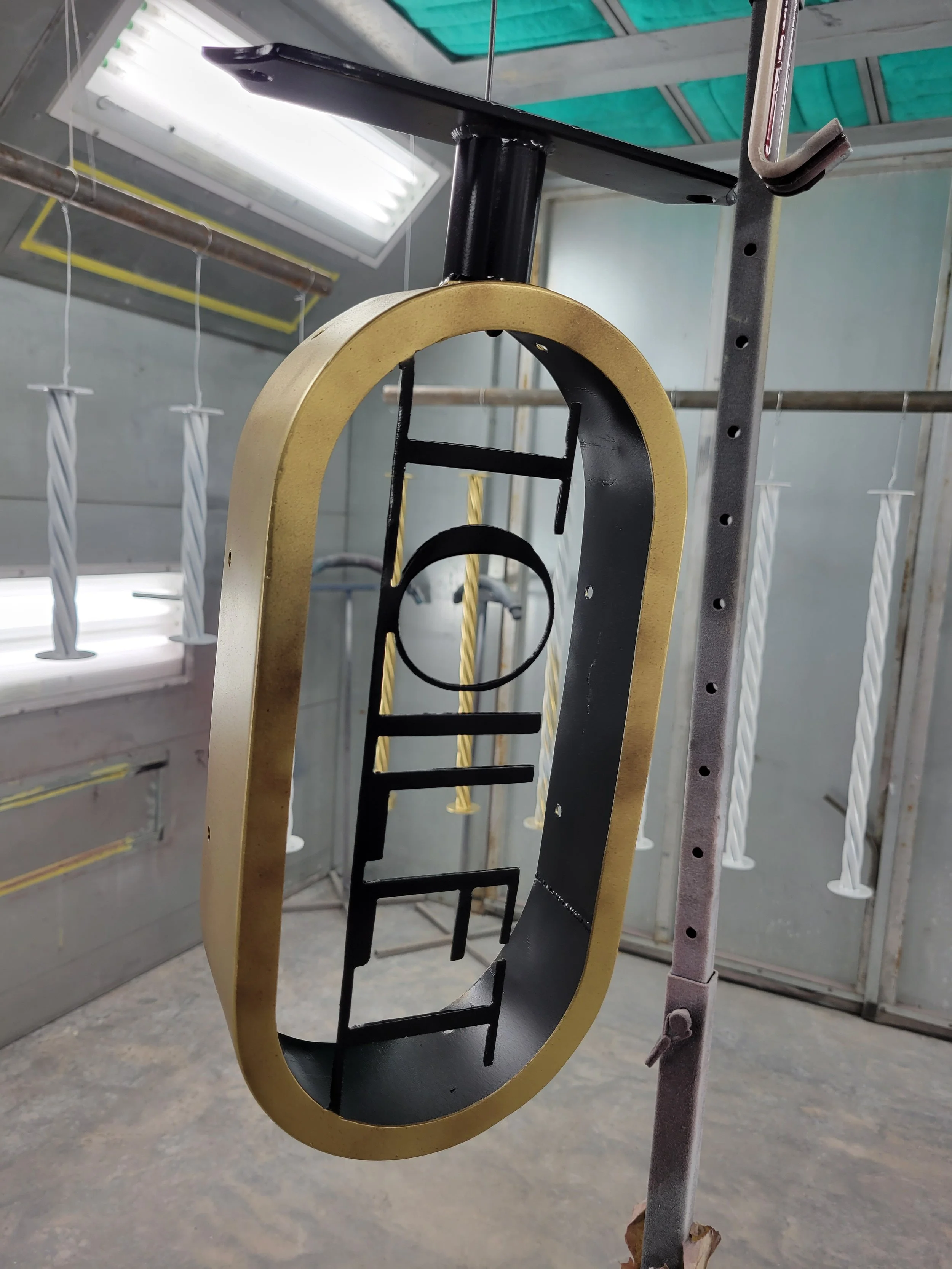 A black metal frame in the shape of numbers with a gold border, hanging in a workshop or garage with unfinished walls and ropes in the background.