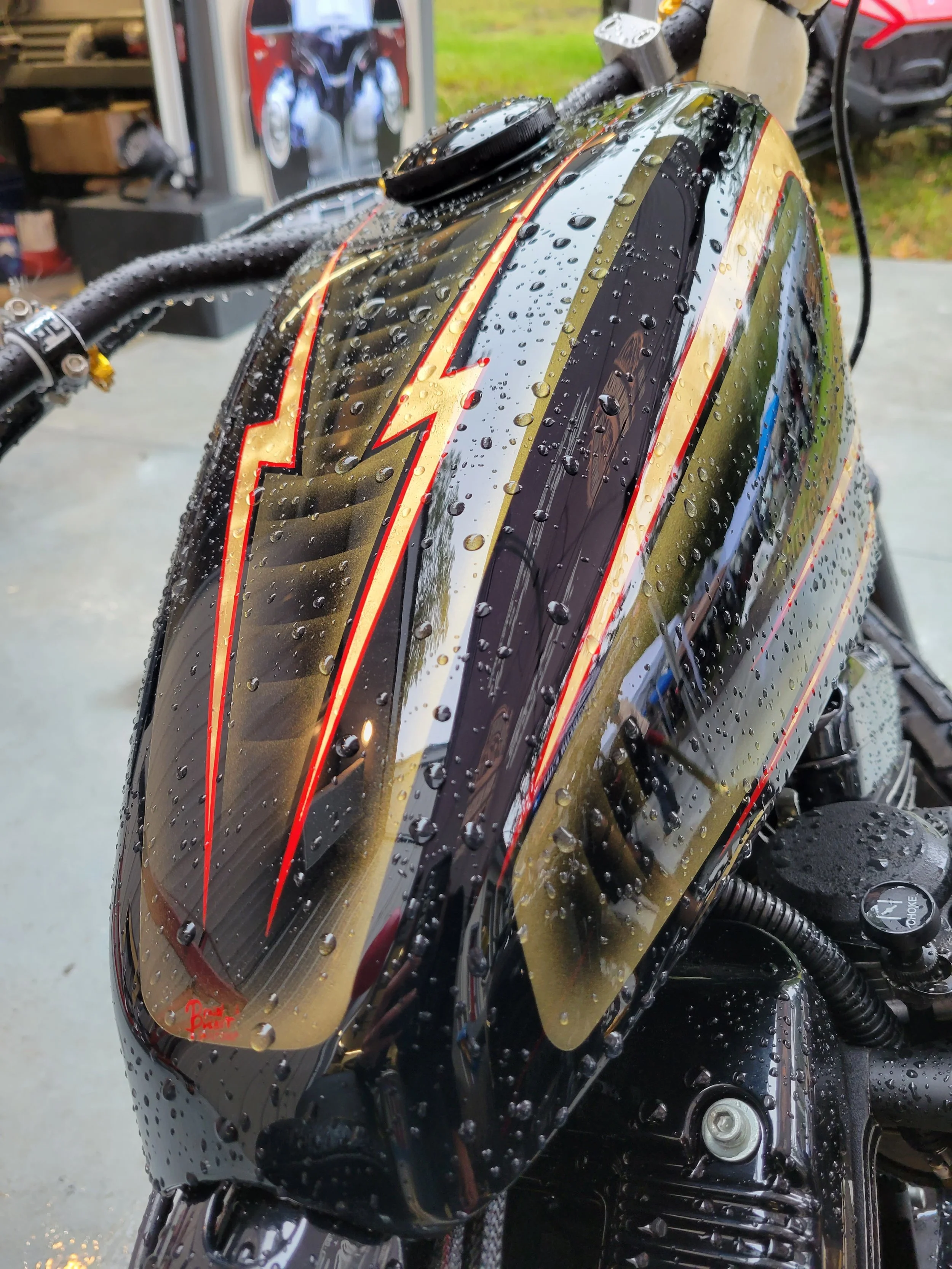 Close-up of a motorcycle fuel tank with water droplets, black with red and gold accents, and a lightning bolt design.