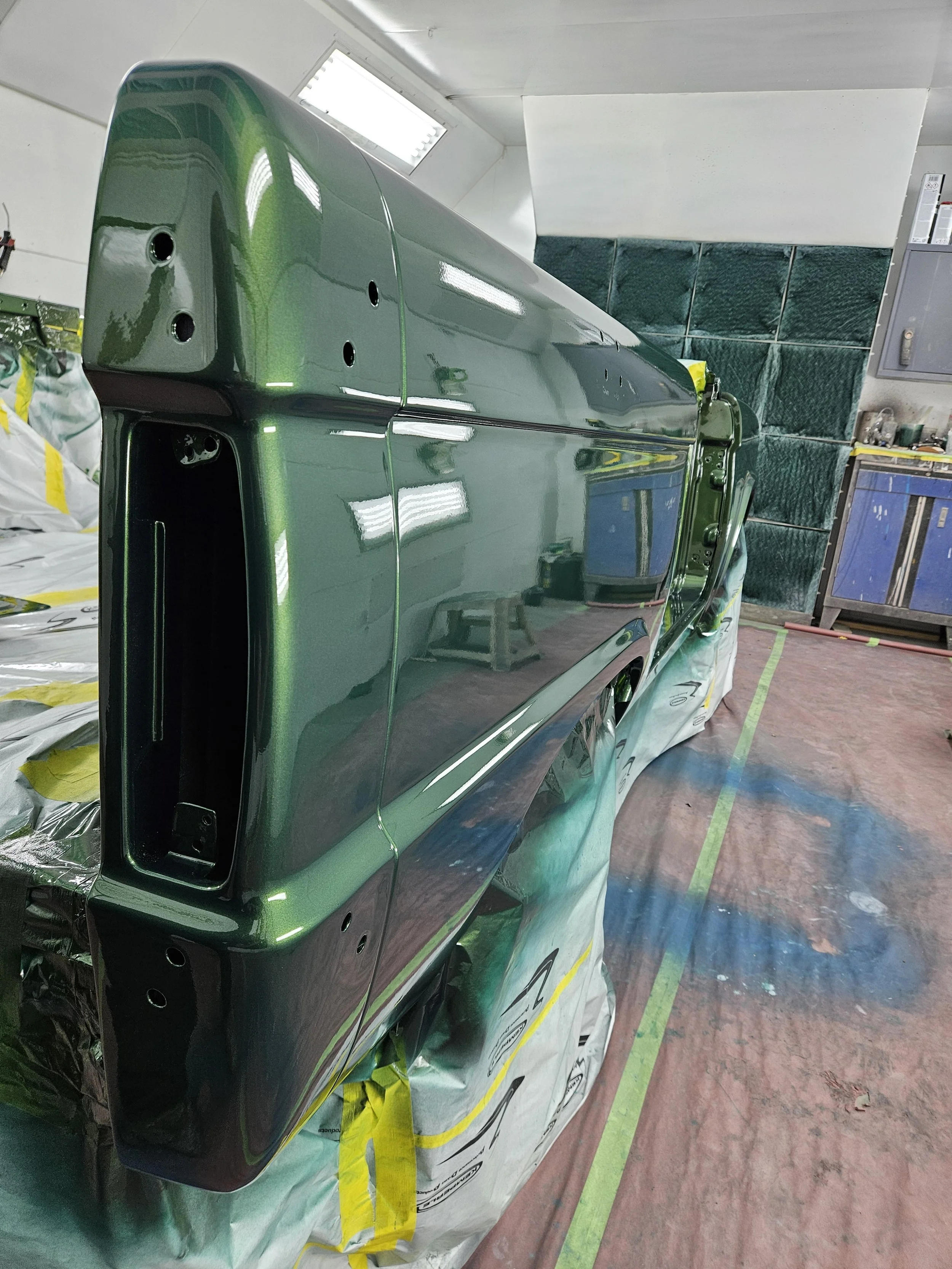 Close-up of a vintage car body painted with a glossy dark green finish in a workshop, with protective masking near the bottom and reflection of workshop lights on its surface.