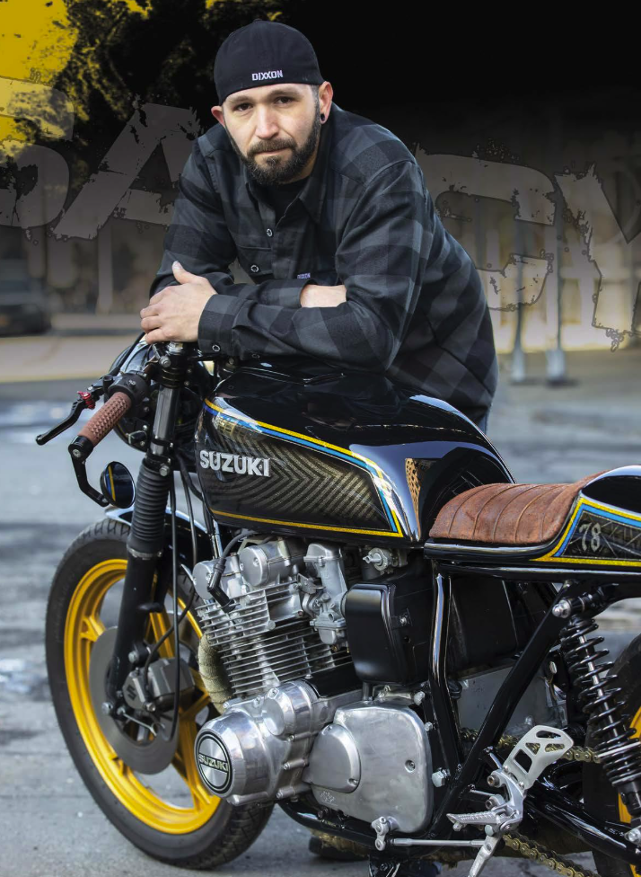 A man with a beard wearing a black cap and black plaid shirt leaning on a customized Suzuki motorcycle in an outdoor parking lot.