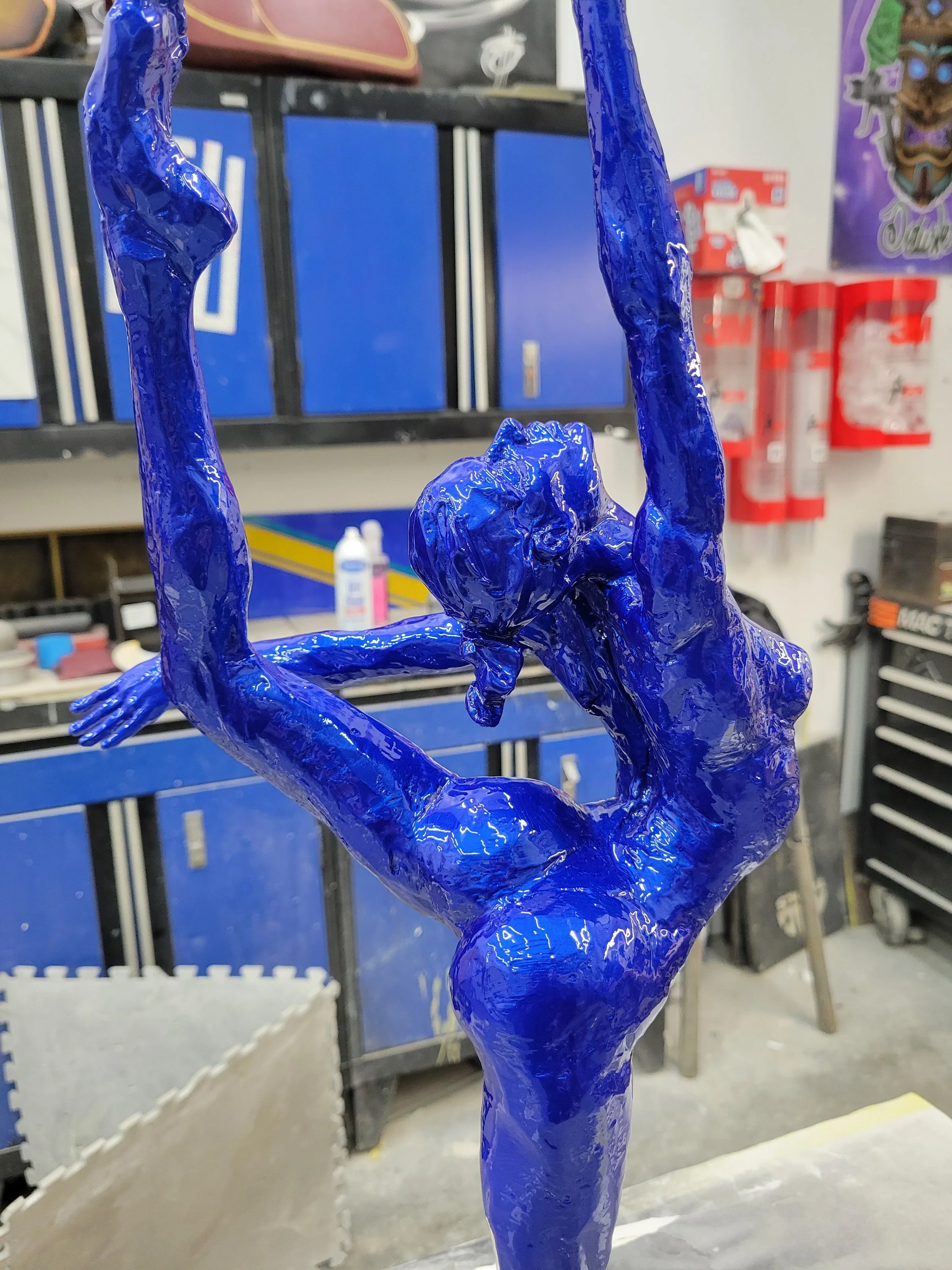 Blue sculpture of a human figure in a dynamic pose, hanging upside down with arms extended, in an artist's workshop.