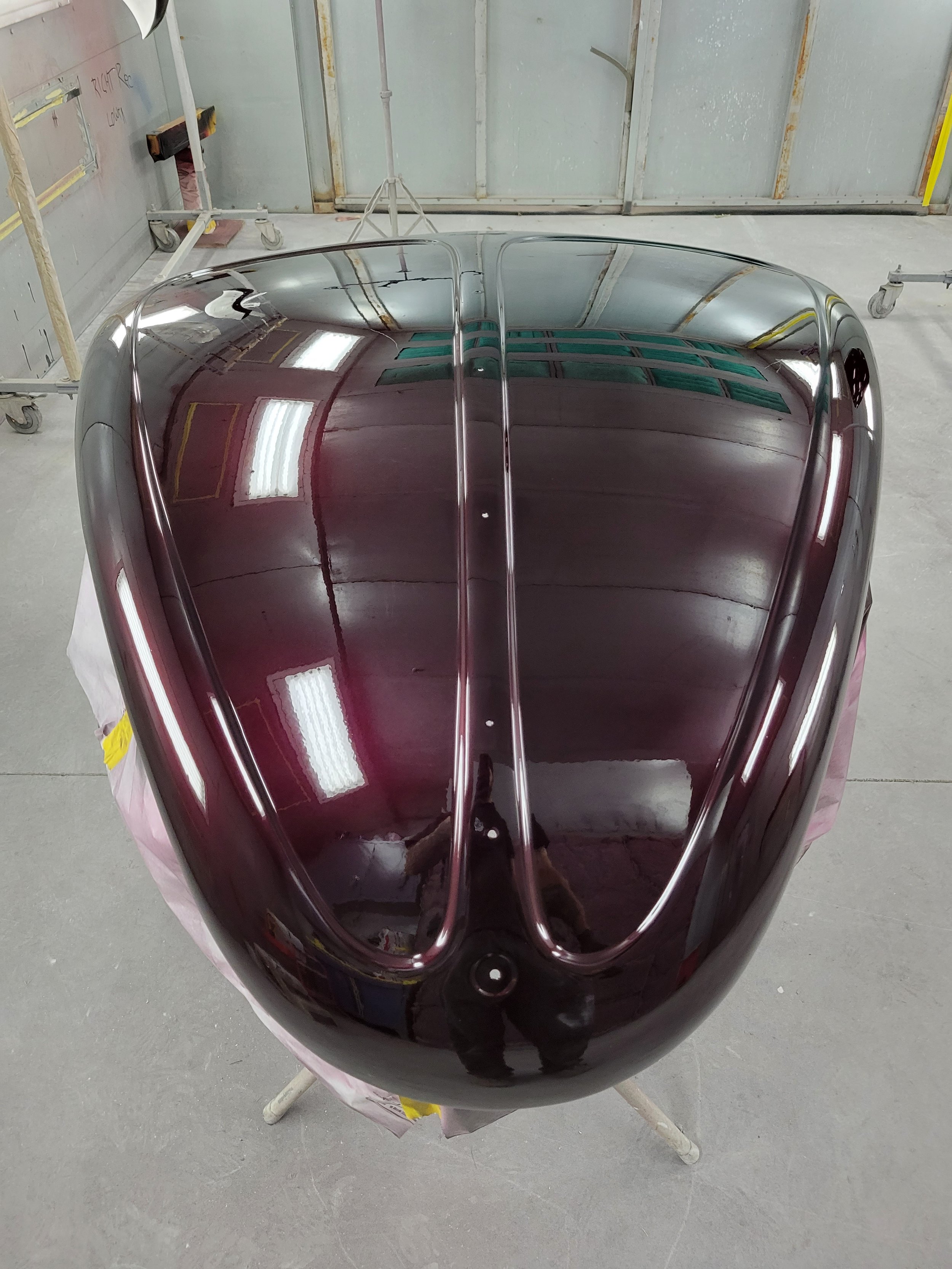 A shiny, dark-colored car hood with a sleek, aerodynamic design, reflecting the overhead lights and surroundings, in a garage or workshop setting.