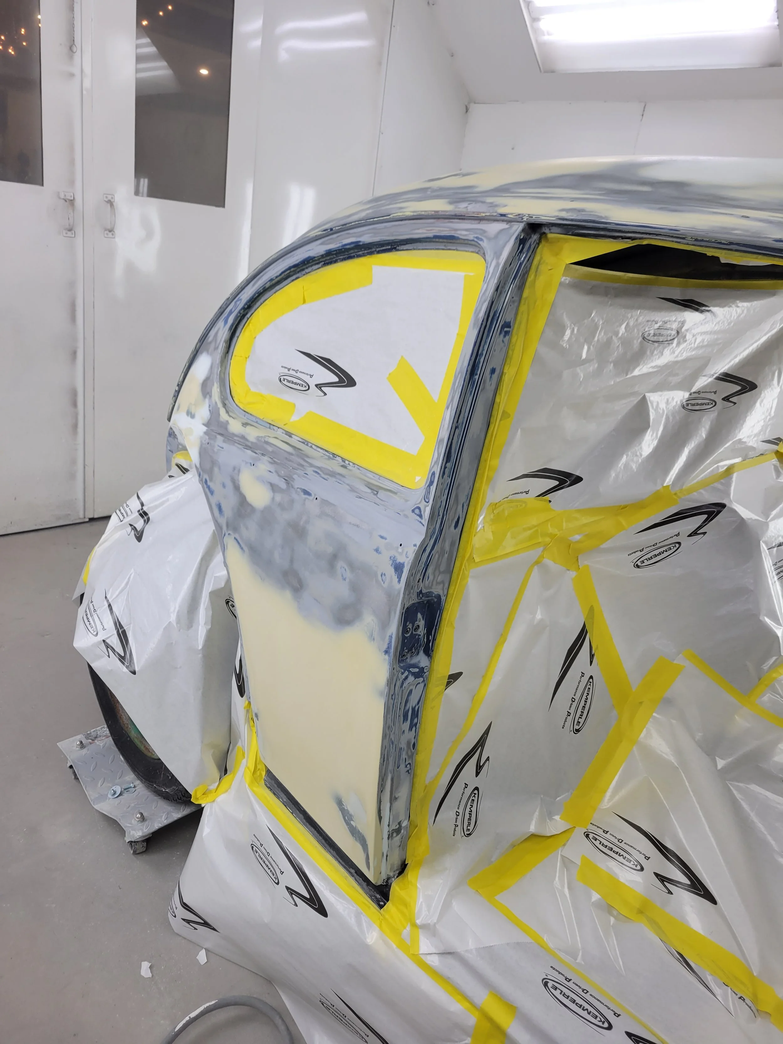 A car in a paint booth being prepared for painting, with masking tape and paper covering parts of the vehicle.