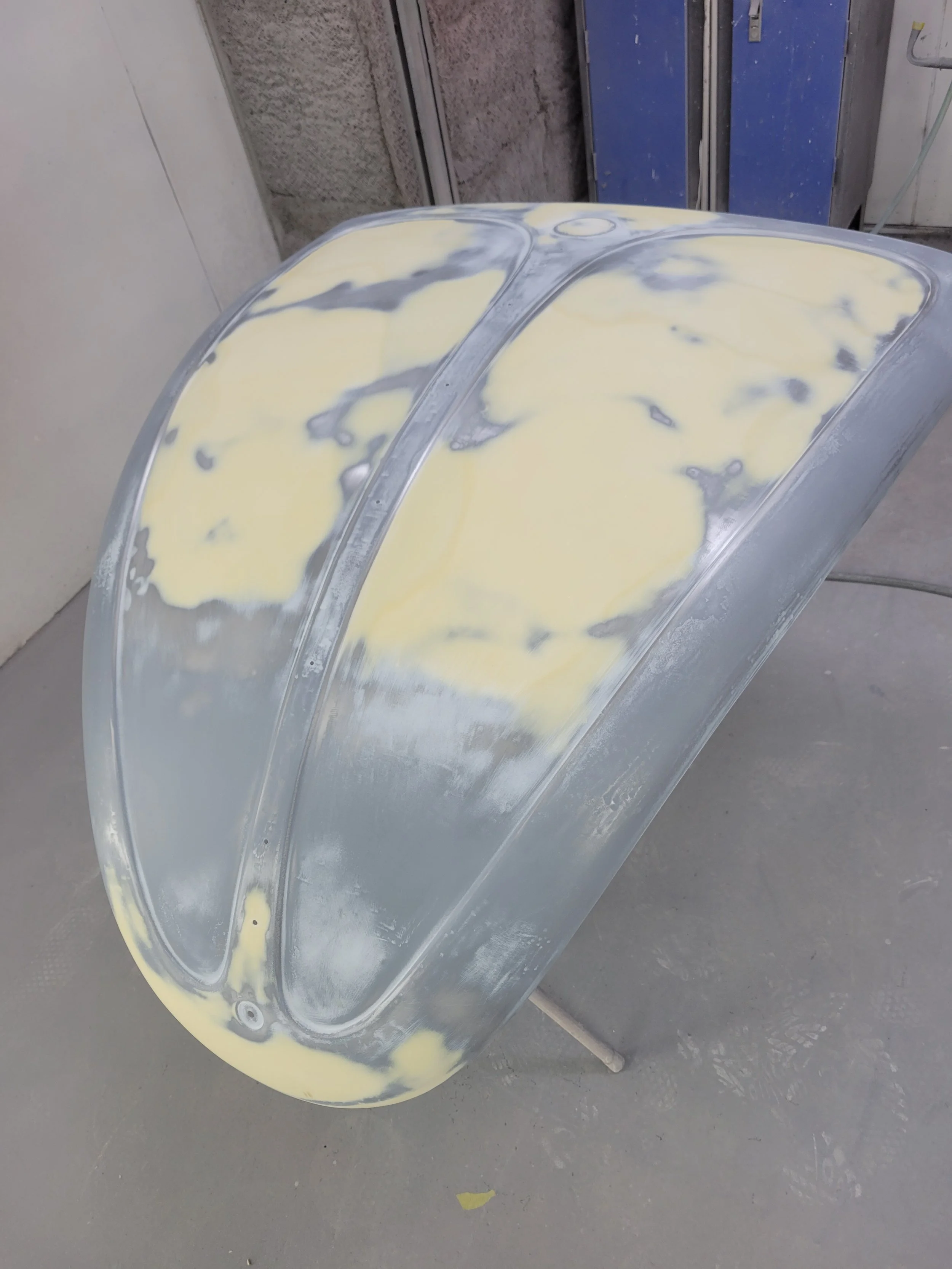 Unpainted car hood with primer and some yellow filler spots, resting on a stand in a workshop.