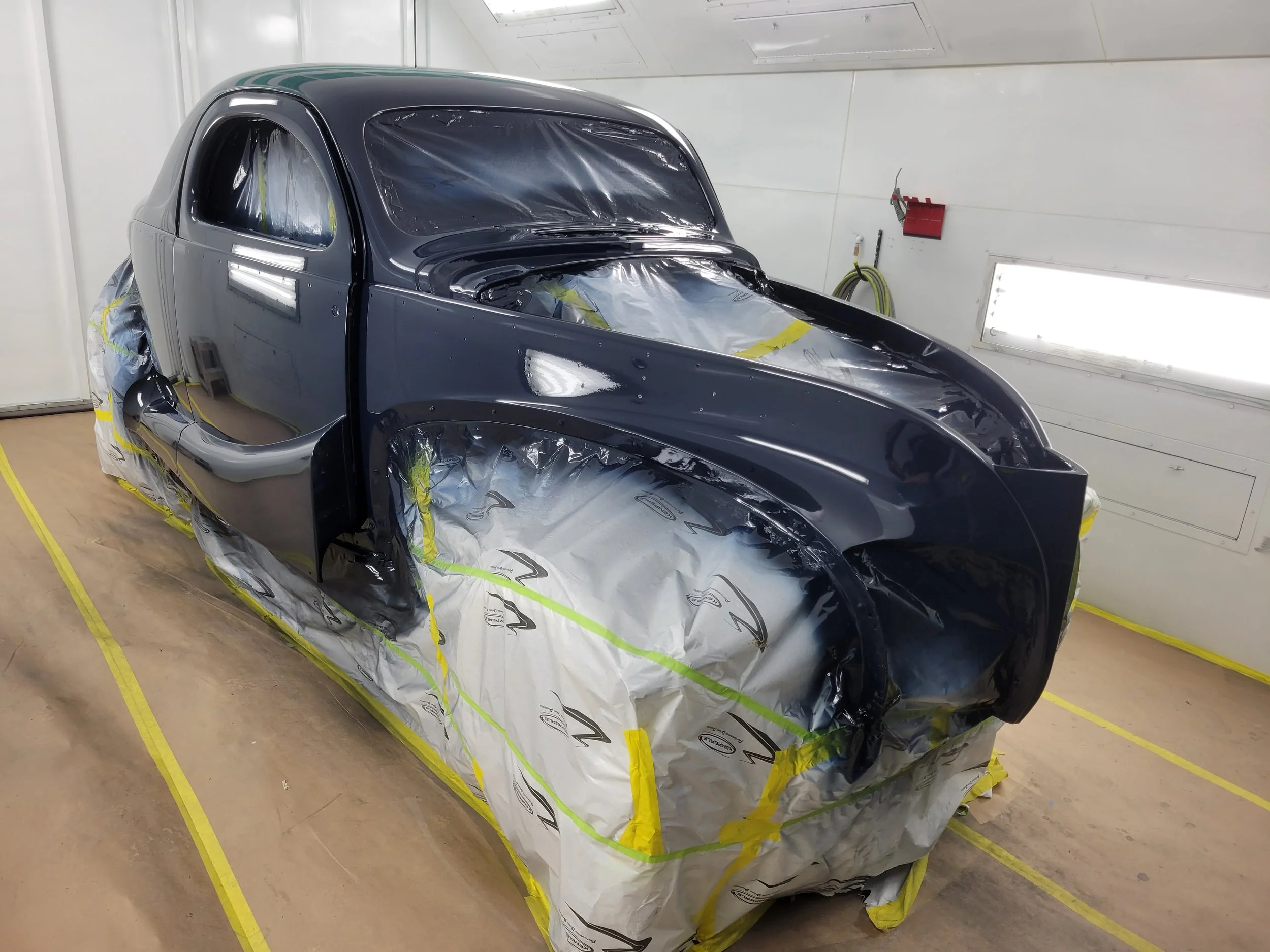 A black car in a paint booth, masked for painting, with plastic and tape covering parts of the car, ready for painting or clear coat application.