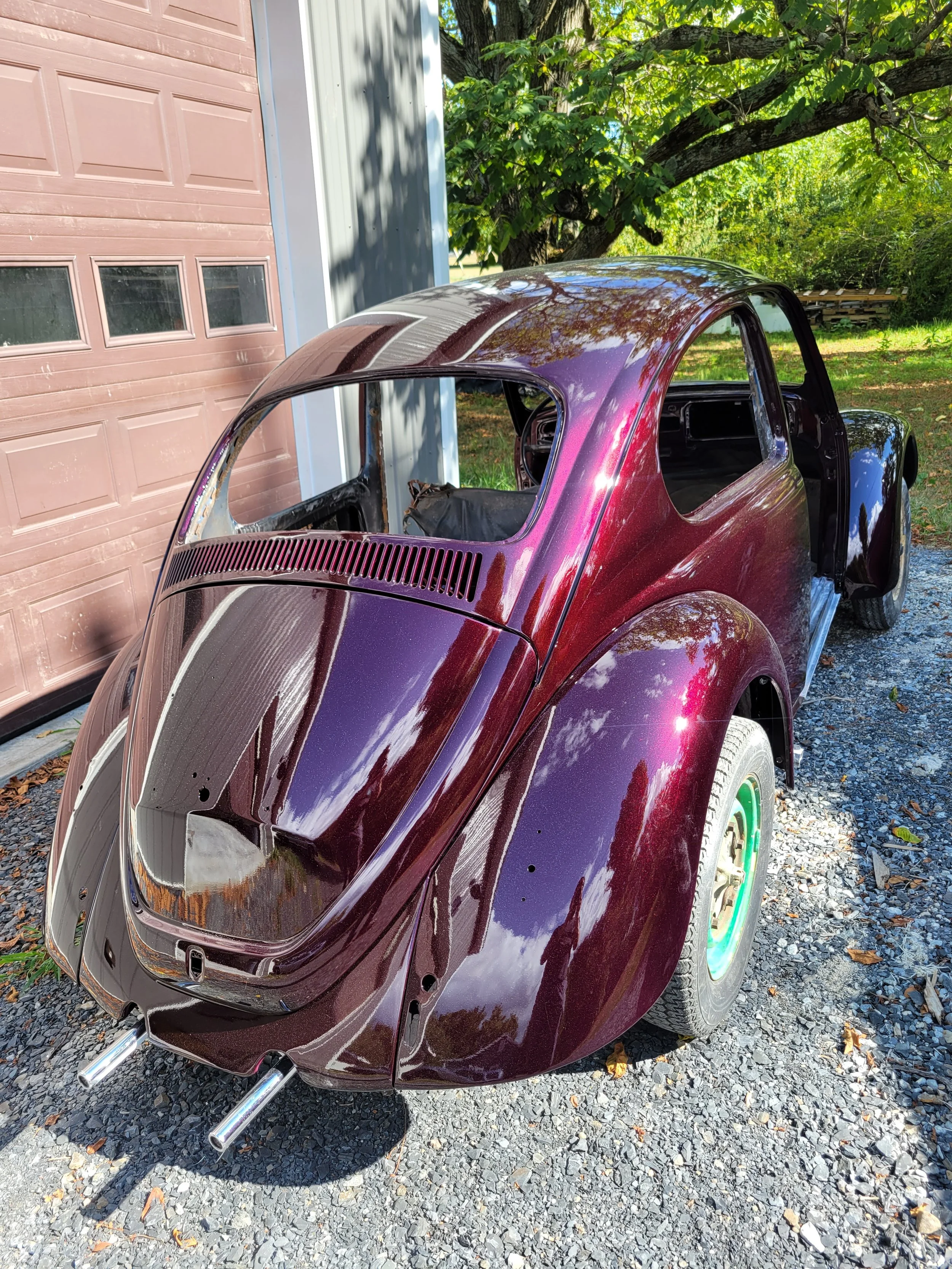 A vintage car body painted in a shiny, multicolored purple and burgundy finish, parked outside on a gravel surface near a garage, with trees and greenery in the background.