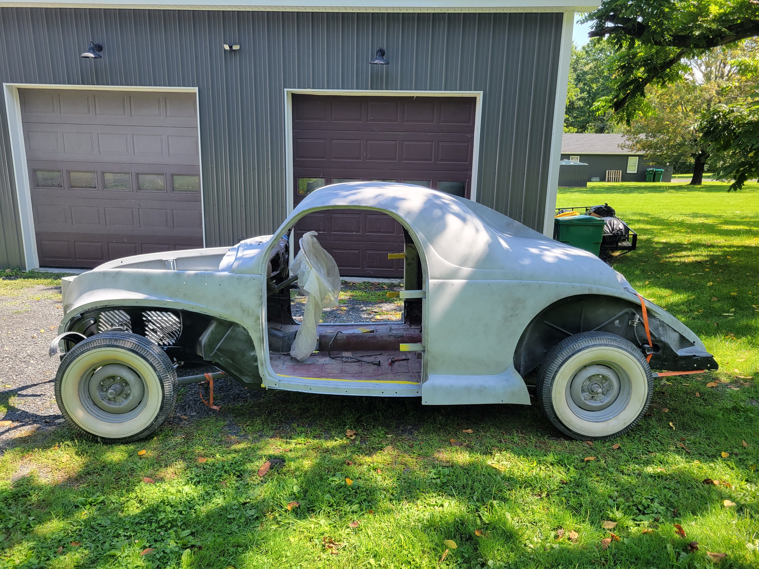 An unfinished vintage car body shell on grass in front of a garage, with a tree and yard in the background.