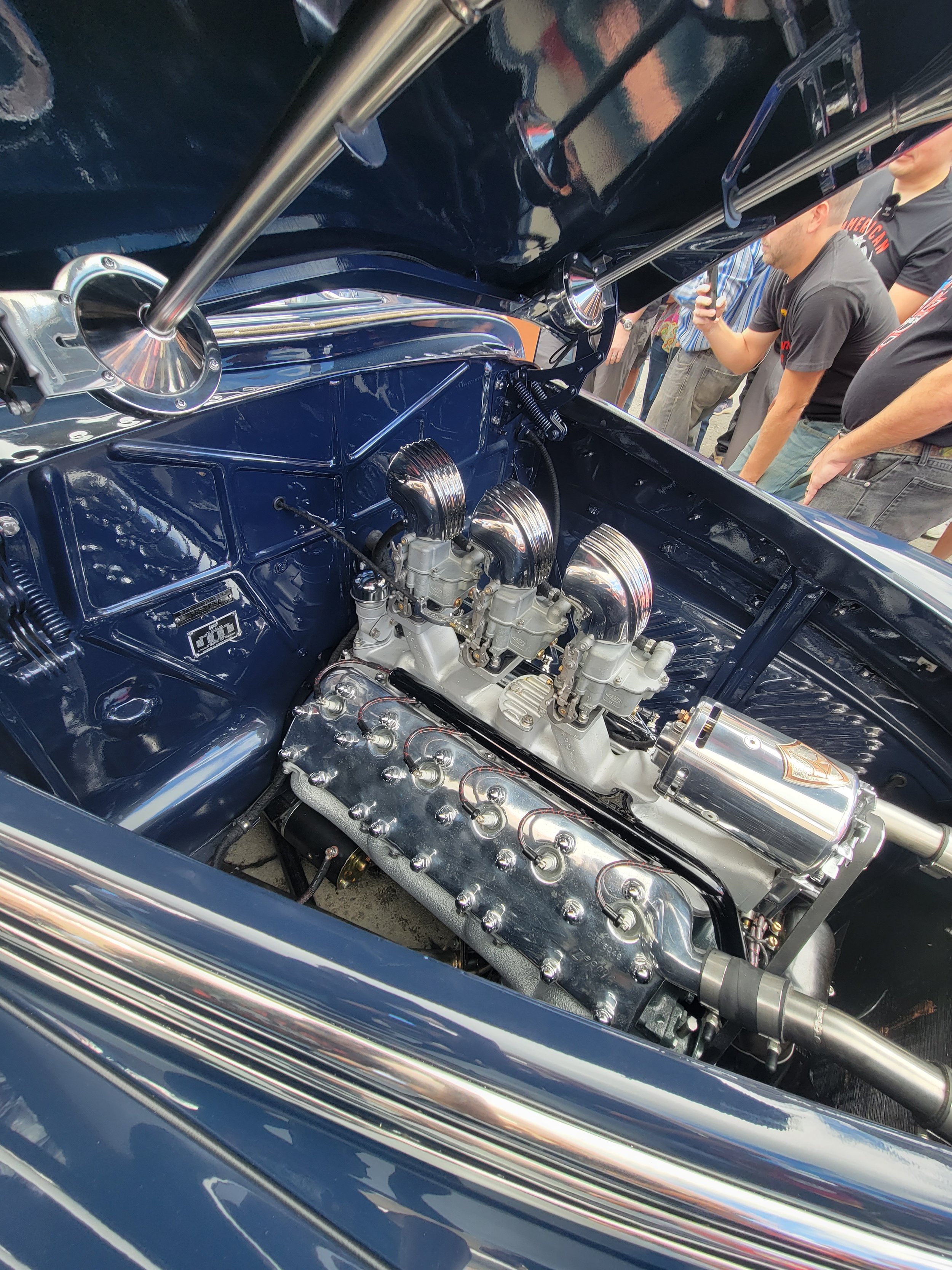Inside the engine compartment of a vintage car, showing polished metallic engine parts and blue-painted interior walls, with a crowd of people in the background.