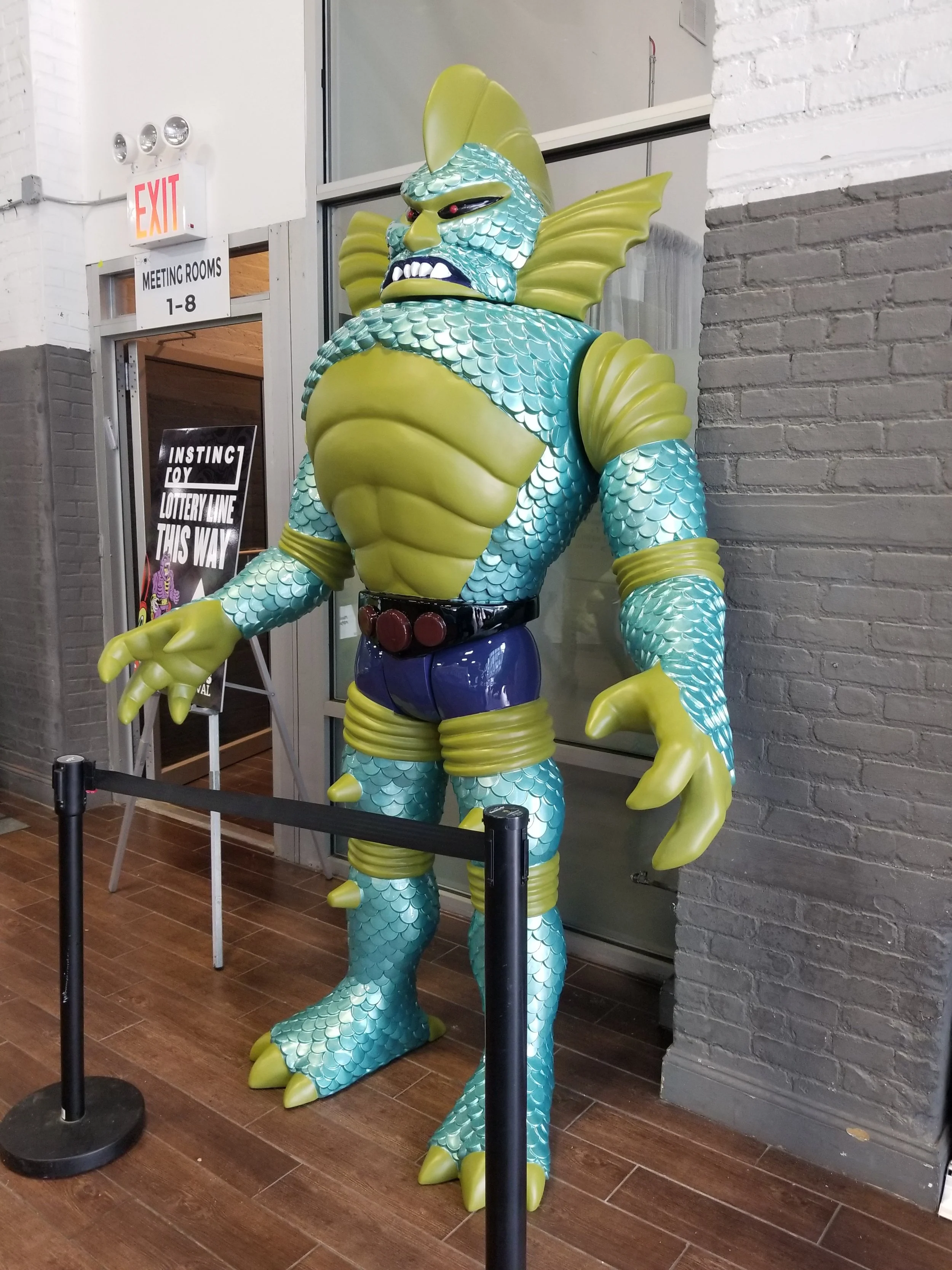 A large cartoonish statue of a humanoid sea creature with fish scales, fins, and claws, dressed in blue shorts and a belt, positioned inside a building.