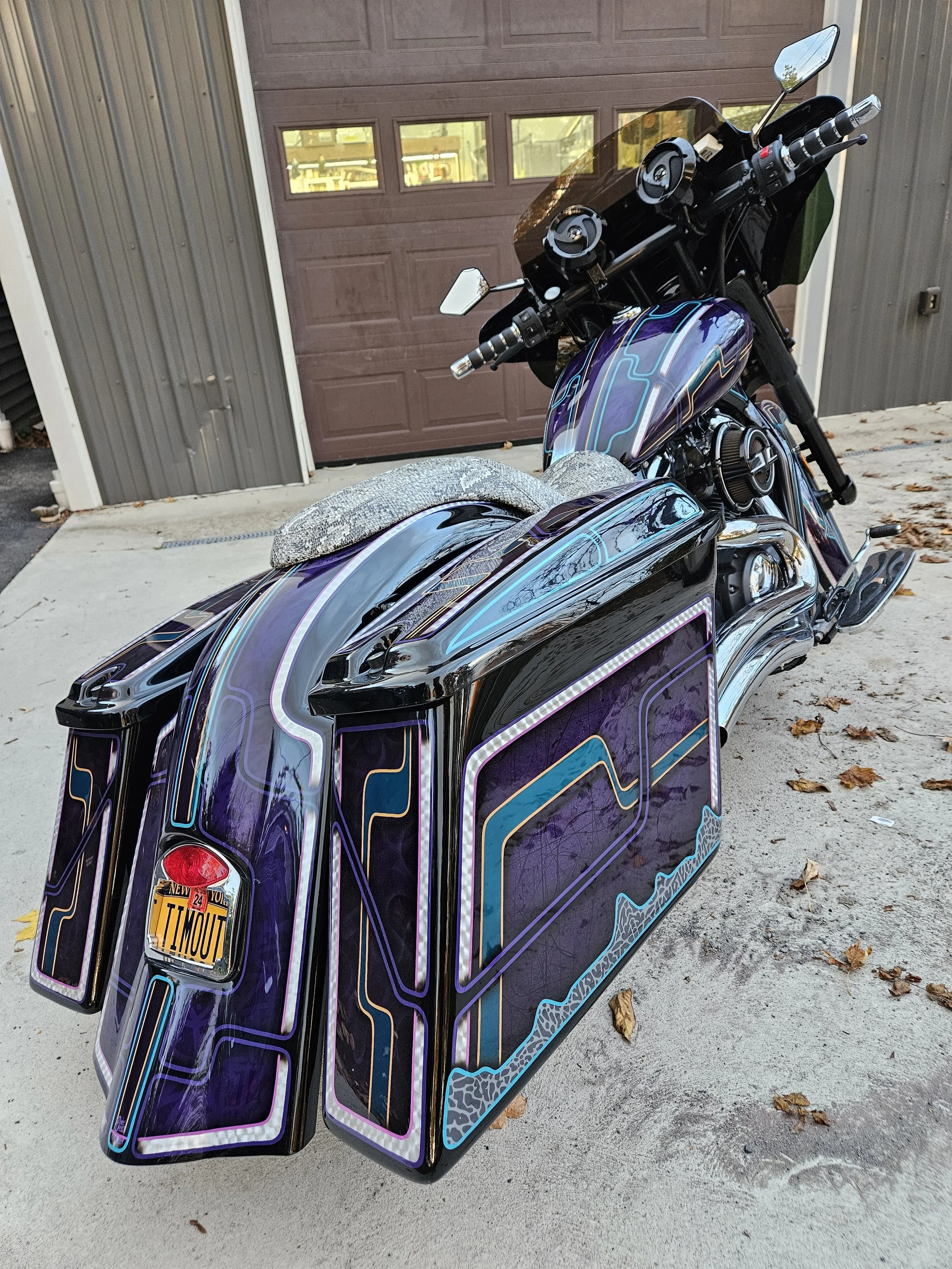 A motorcycle with neon purple and blue custom paint, featuring a rear trunk and saddlebags, parked on a concrete driveway in front of a brown garage door and gray siding house.