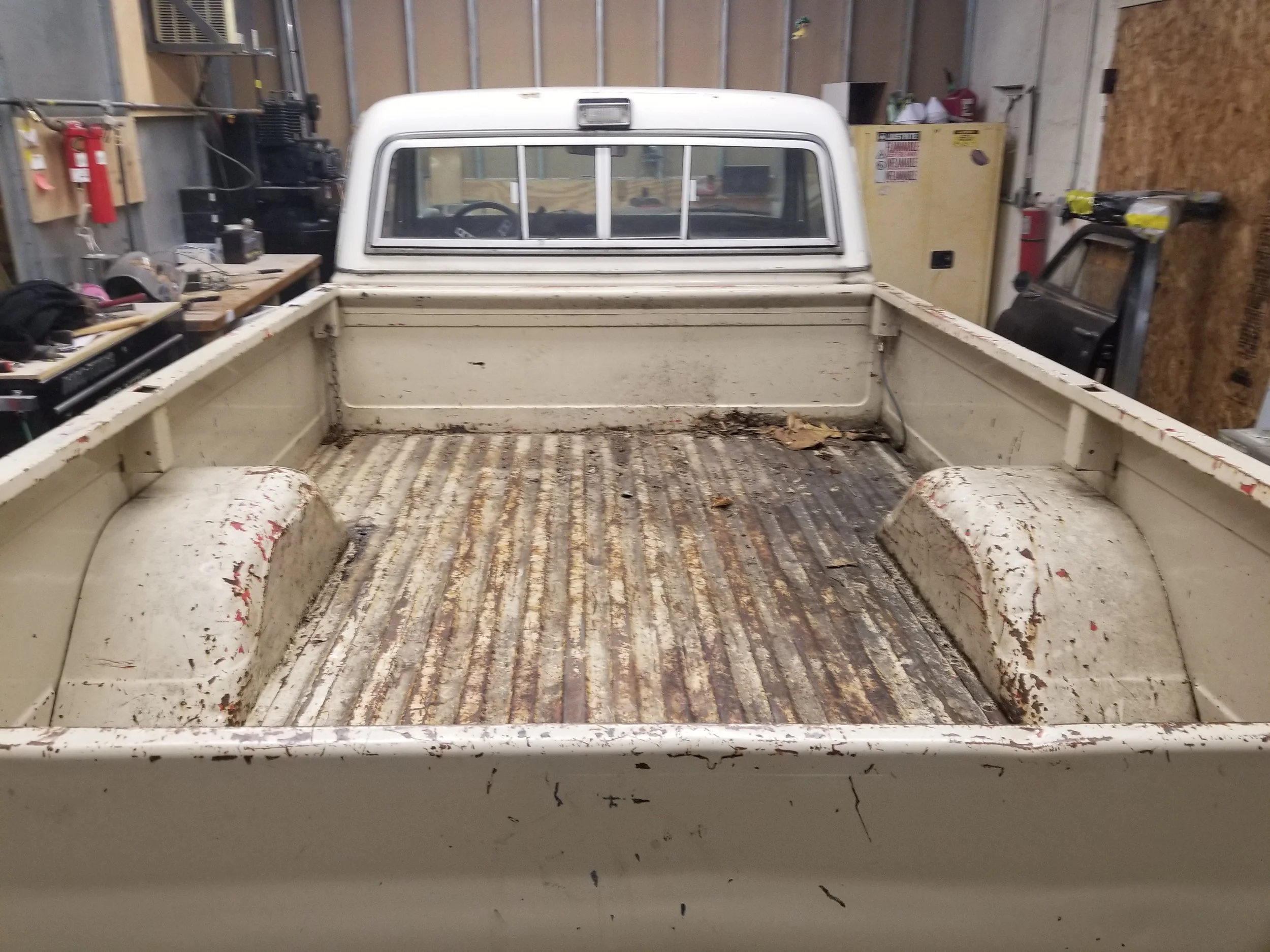 The bed of an old white pickup truck with rust and debris, parked inside a garage or workshop.