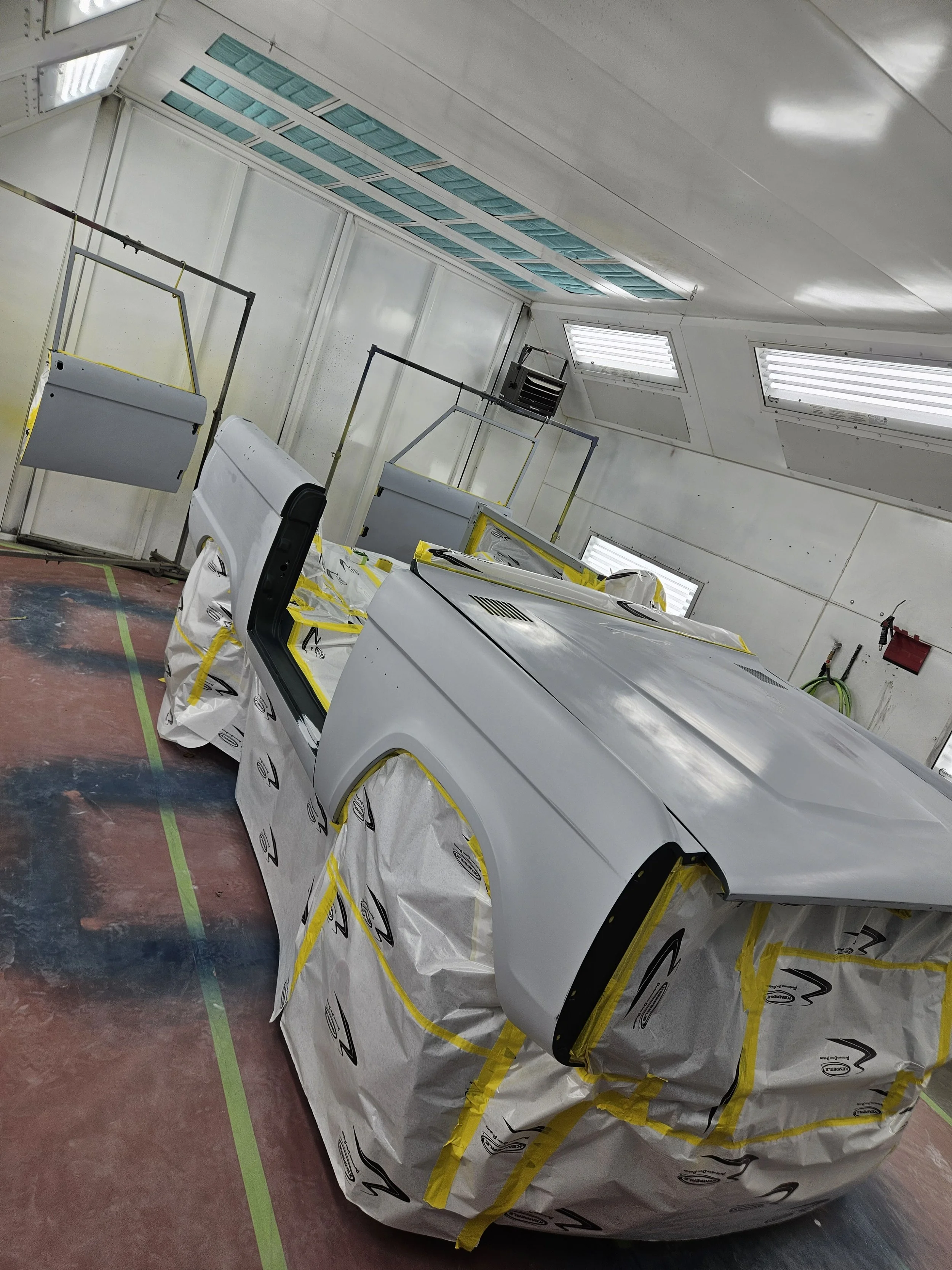 Auto body painted white in a paint booth, covered with protective paper and tape, with some parts masked for painting.