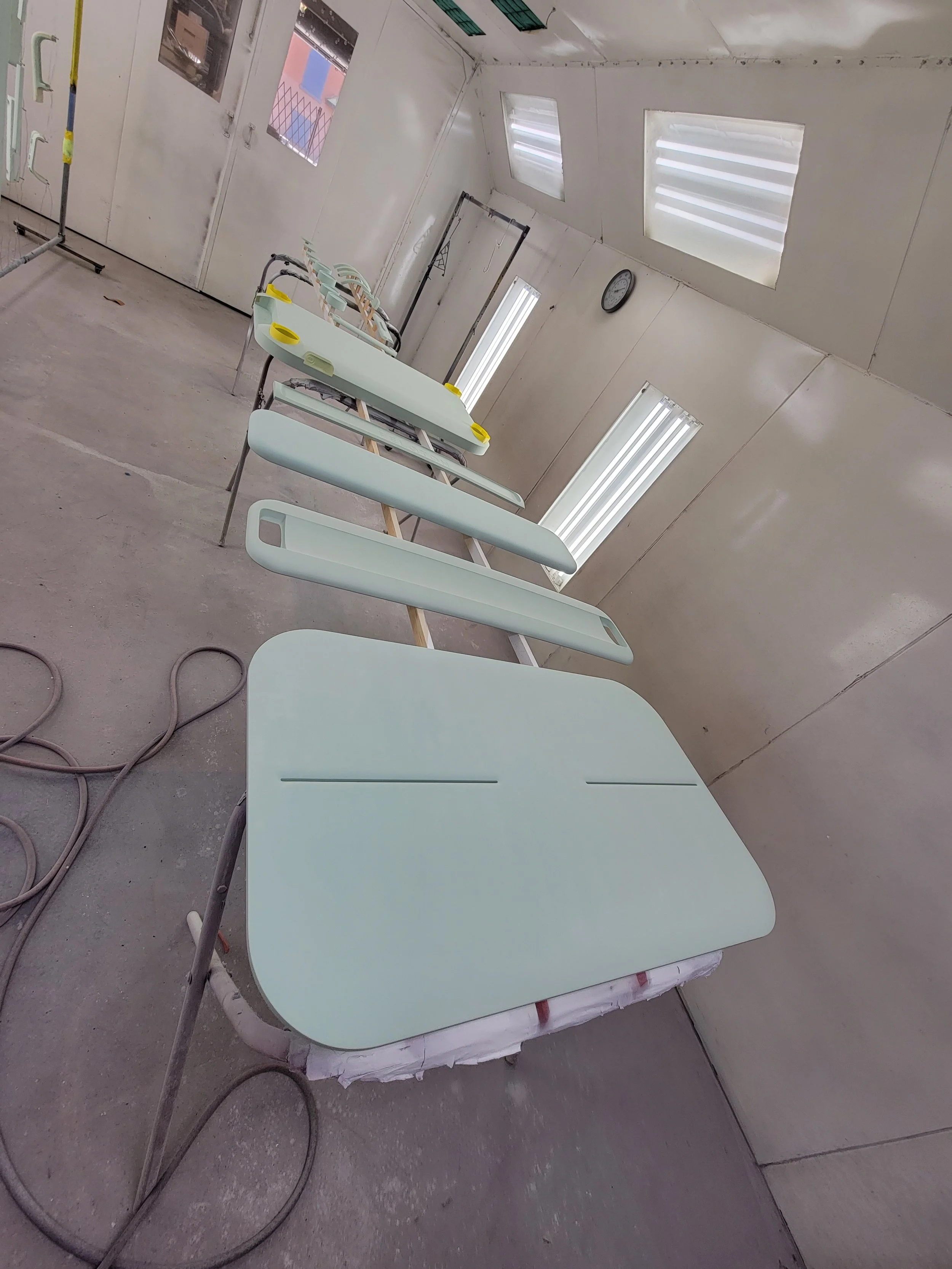 A row of painted hospital bed frames in a workshop or paint booth, with some beds partially assembled. The room has white walls, fluorescent lighting, a clock on the wall, and windows with blinds.