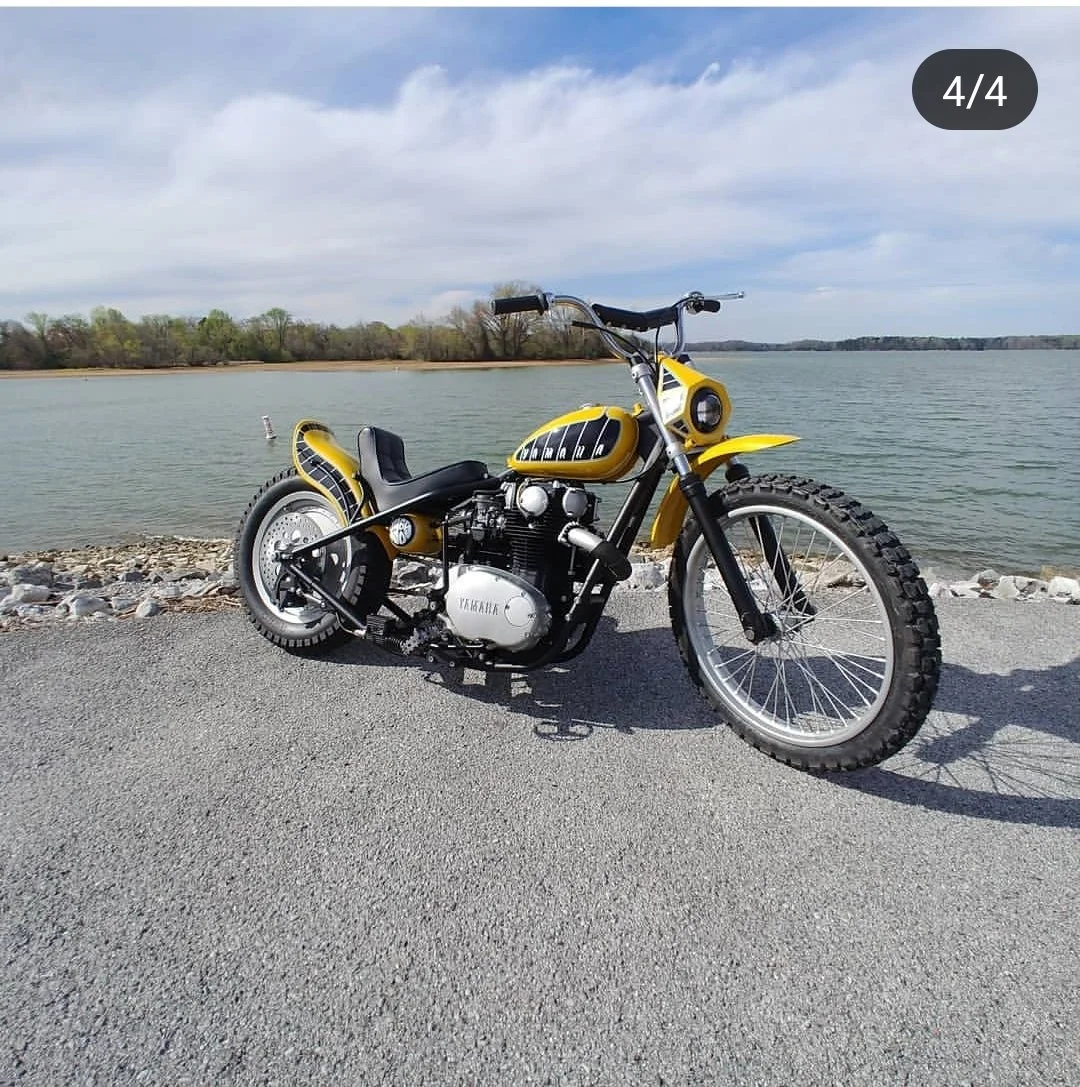 Yellow and black vintage motorcycle parked on a paved area by a body of water with trees and a cloudy sky in the background.