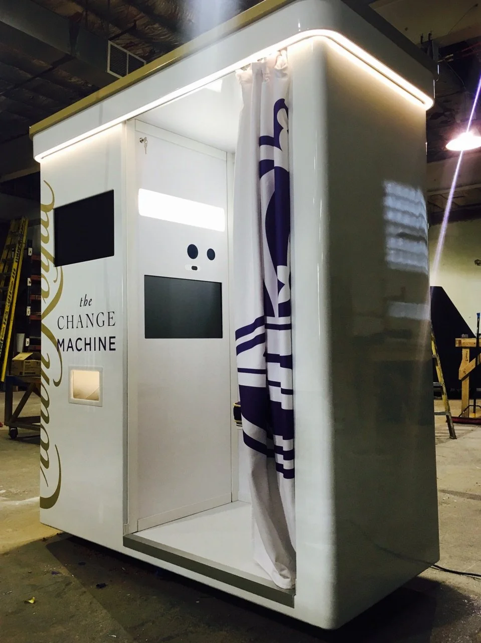 A white mobile photo booth called 'the CHANGE MACHINE' with a curtain inside for photos, two digital screens, and a small slot for prints, located in an indoor setting.