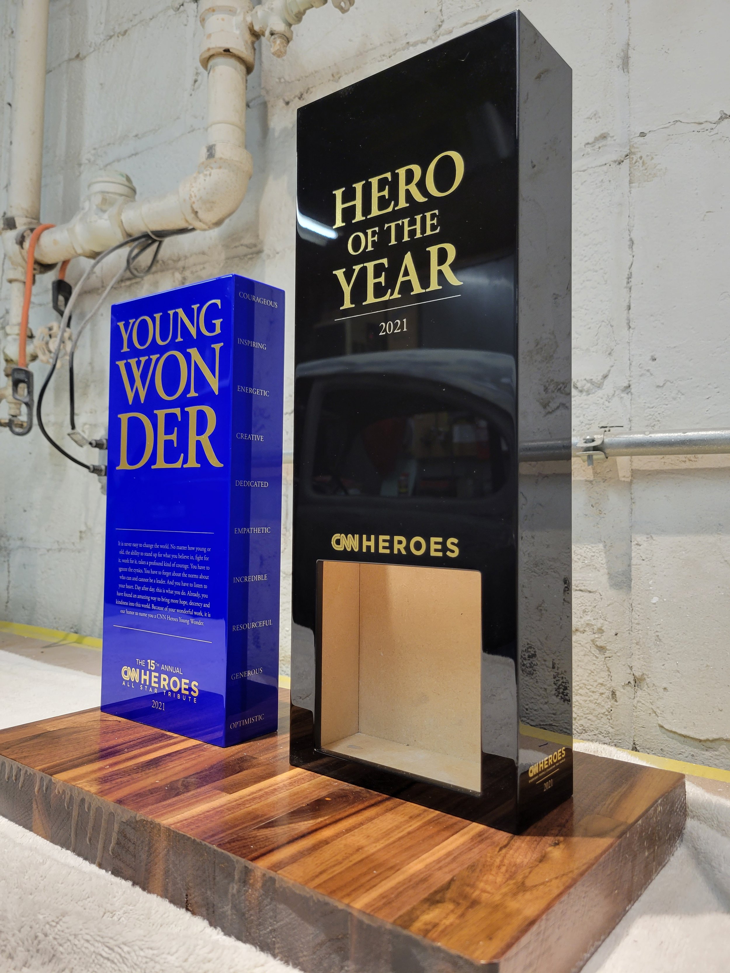 Award trophies for CNN Heroes 2021, one of which is titled 'Hero of the Year,' and the other celebrates young women, placed on a wooden surface against a wall with exposed pipes.