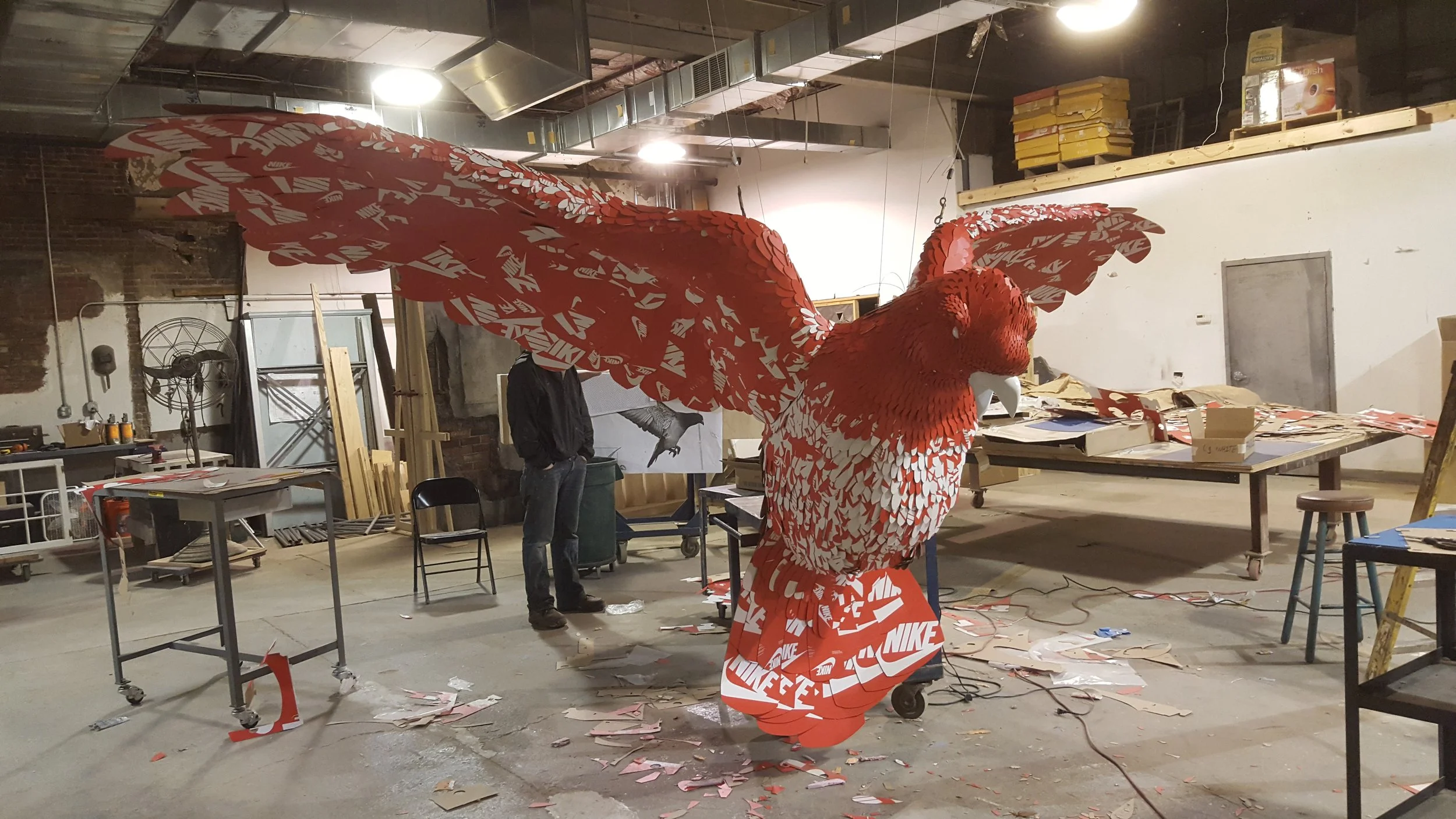 A large, red Nike hoopoe bird sculpture in progress inside an art workshop. The sculpture is made from cut-out Nike logos, with wings spread wide, and is surrounded by woodworking tables, chairs, and scattered art supplies.