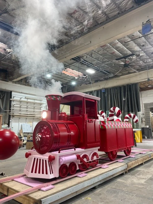 Large decorative red train with white and red candy cane details, emitting smoke, on a wooden platform in a warehouse or workshop.