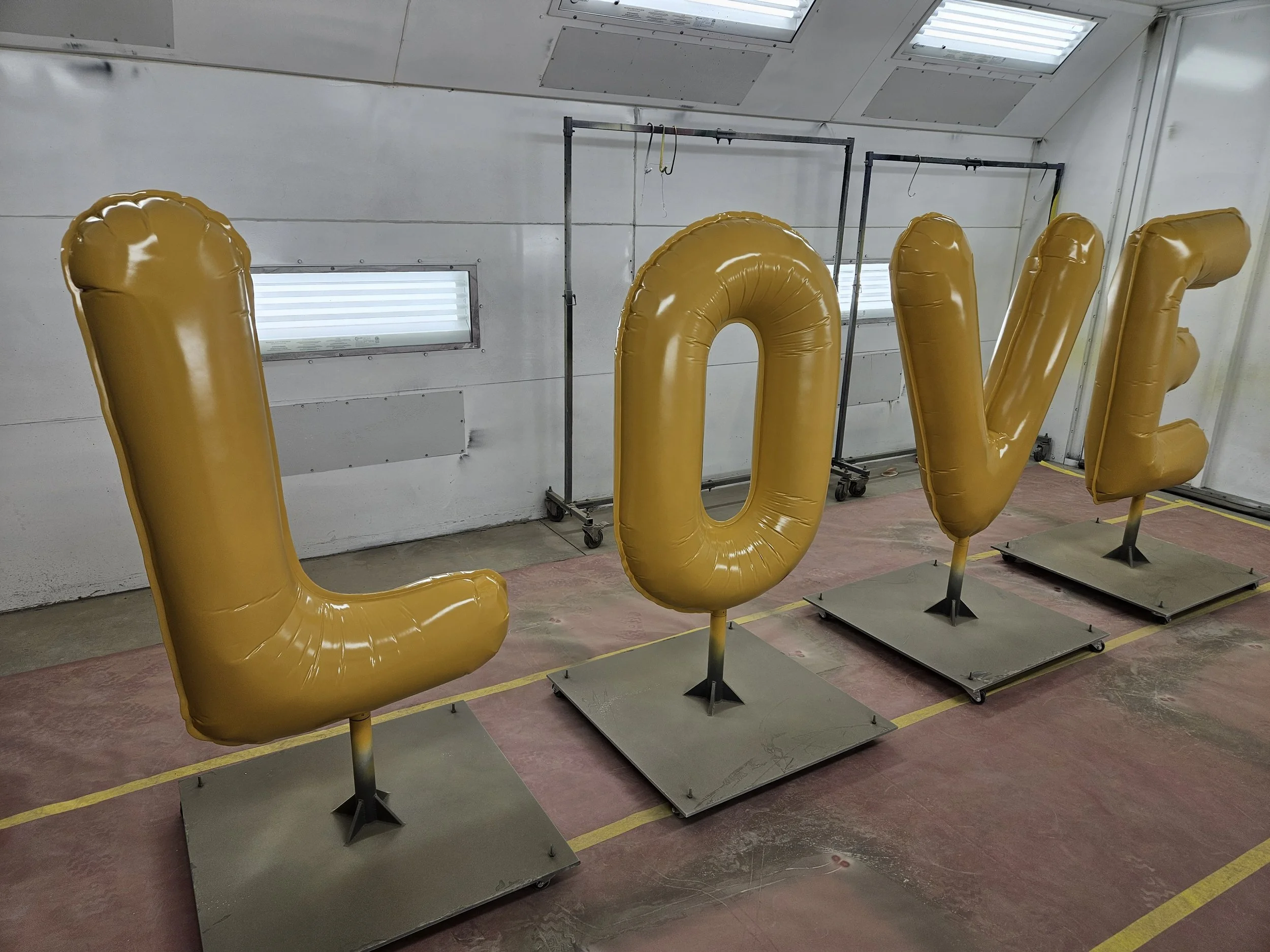 Large gold balloon letters spelling "LOVE" inside a room with white walls, industrial lighting, and a window, mounted on metal bases.