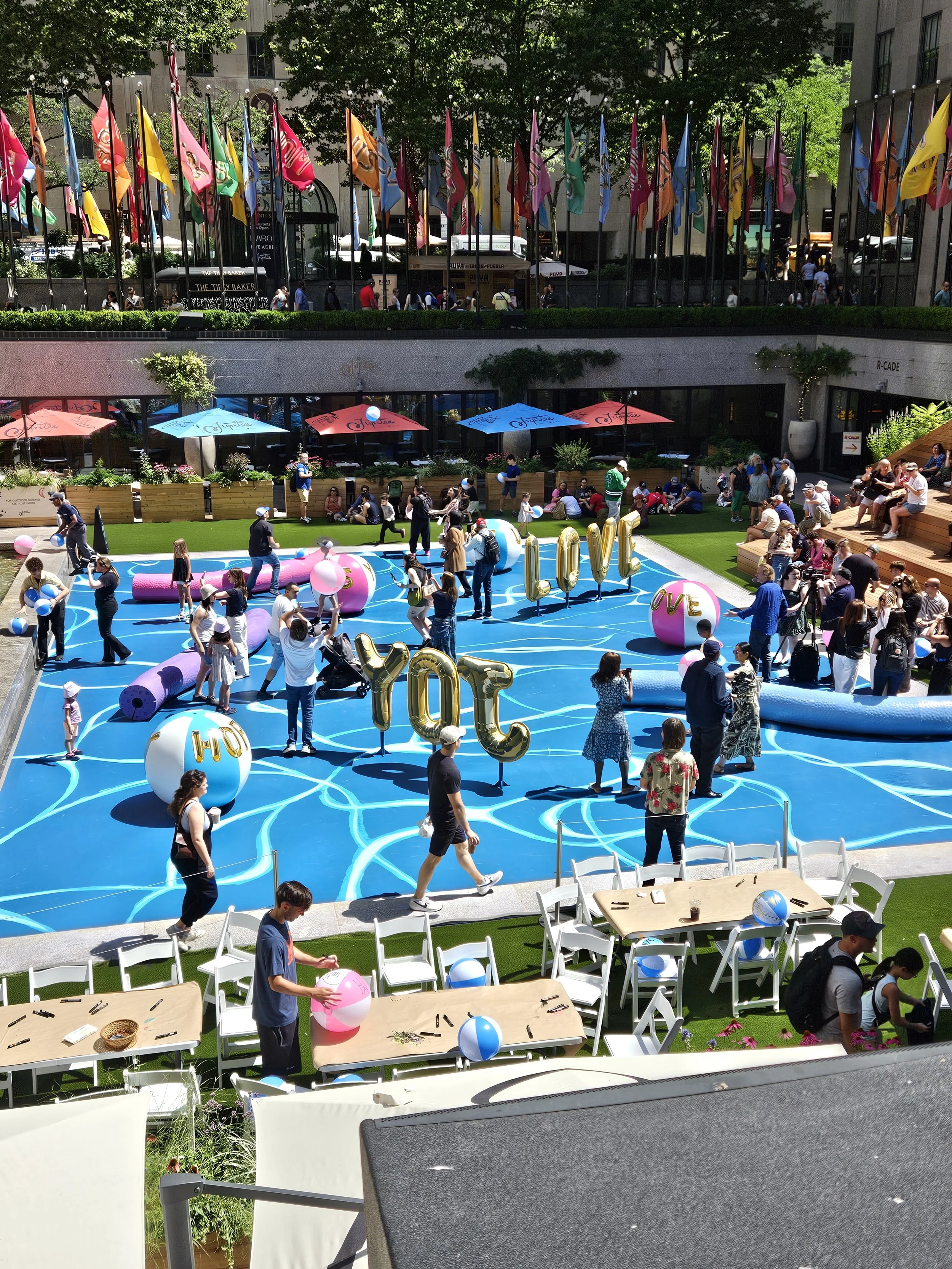 An outdoor celebration in a city square with people gathered around a blue floor with white swirl patterns, holding balloons and taking photos. Large gold helium balloons spell out 'LOVE' and 'YOTC'. There are tables and chairs, some decorated with b