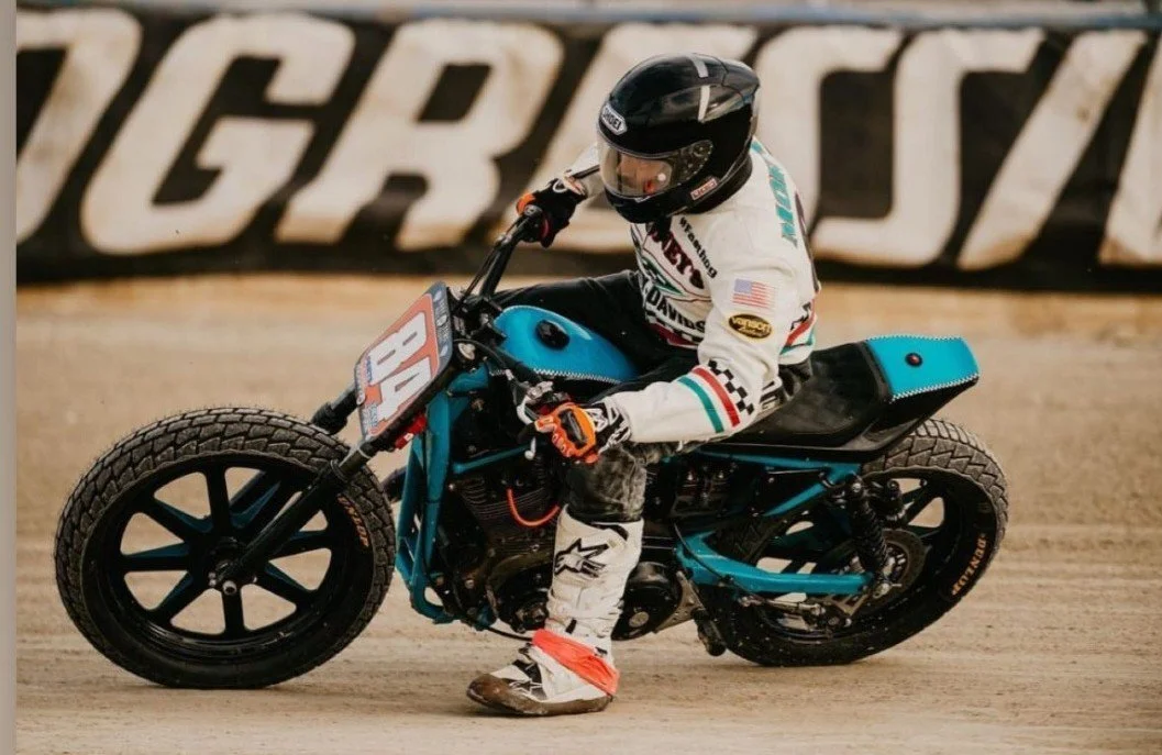 Motorcycle racer in racing gear leaning into a turn on a dirt track, wearing a black helmet and white racing suit with colorful accents.