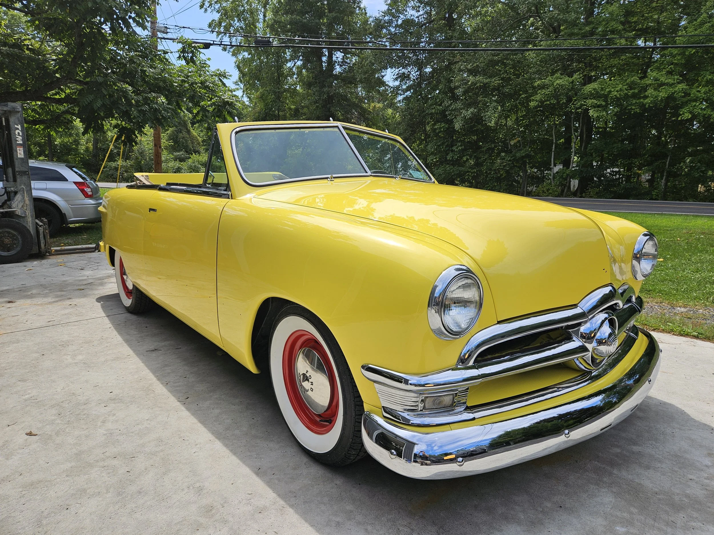 Yellow vintage convertible car parked on a concrete driveway, with a silver SUV and trees in the background.