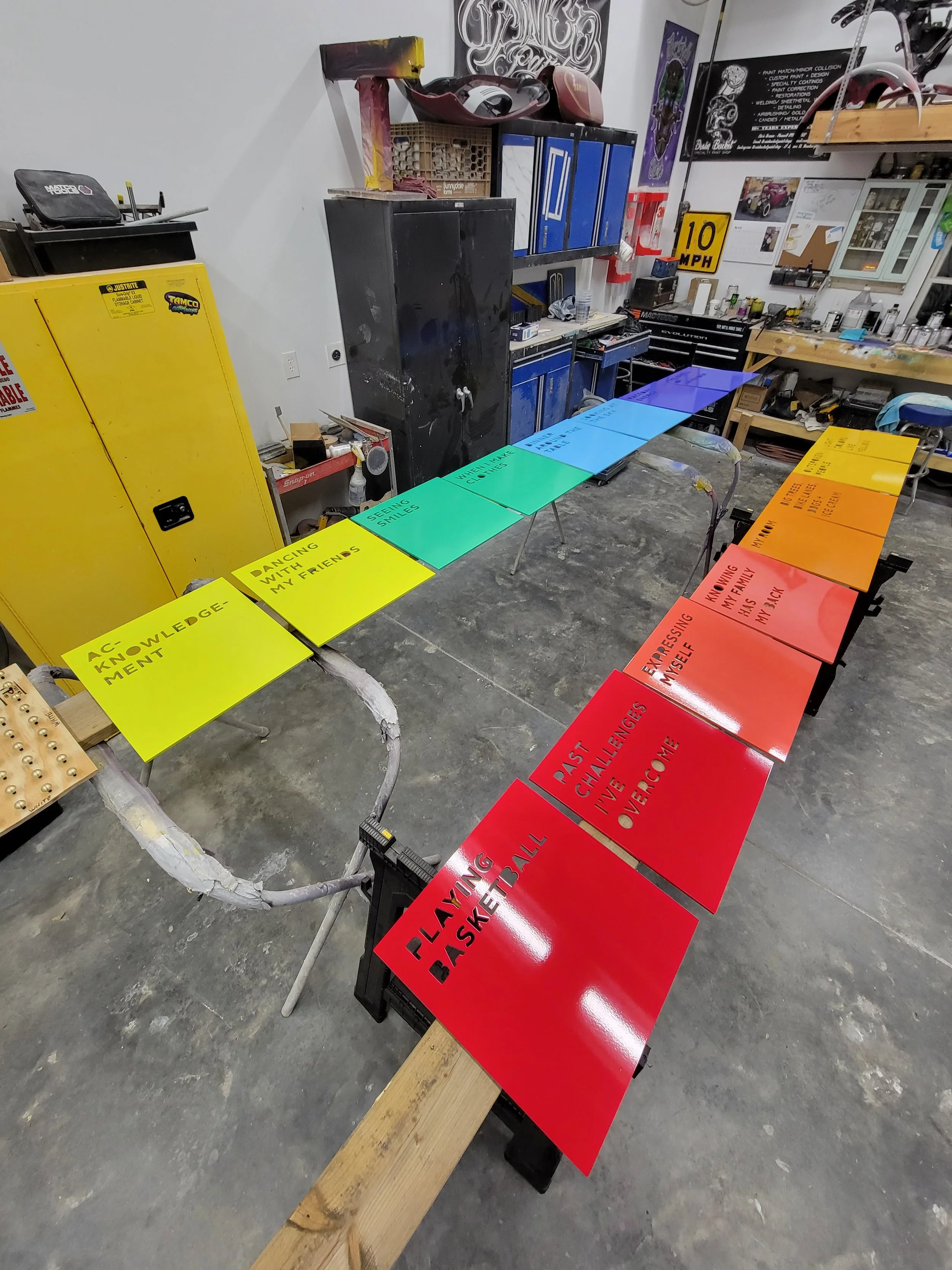 Colorful motivational signs arranged on a workbench in a workshop. The signs are in yellow, green, blue, orange, and red, each with different inspirational phrases.