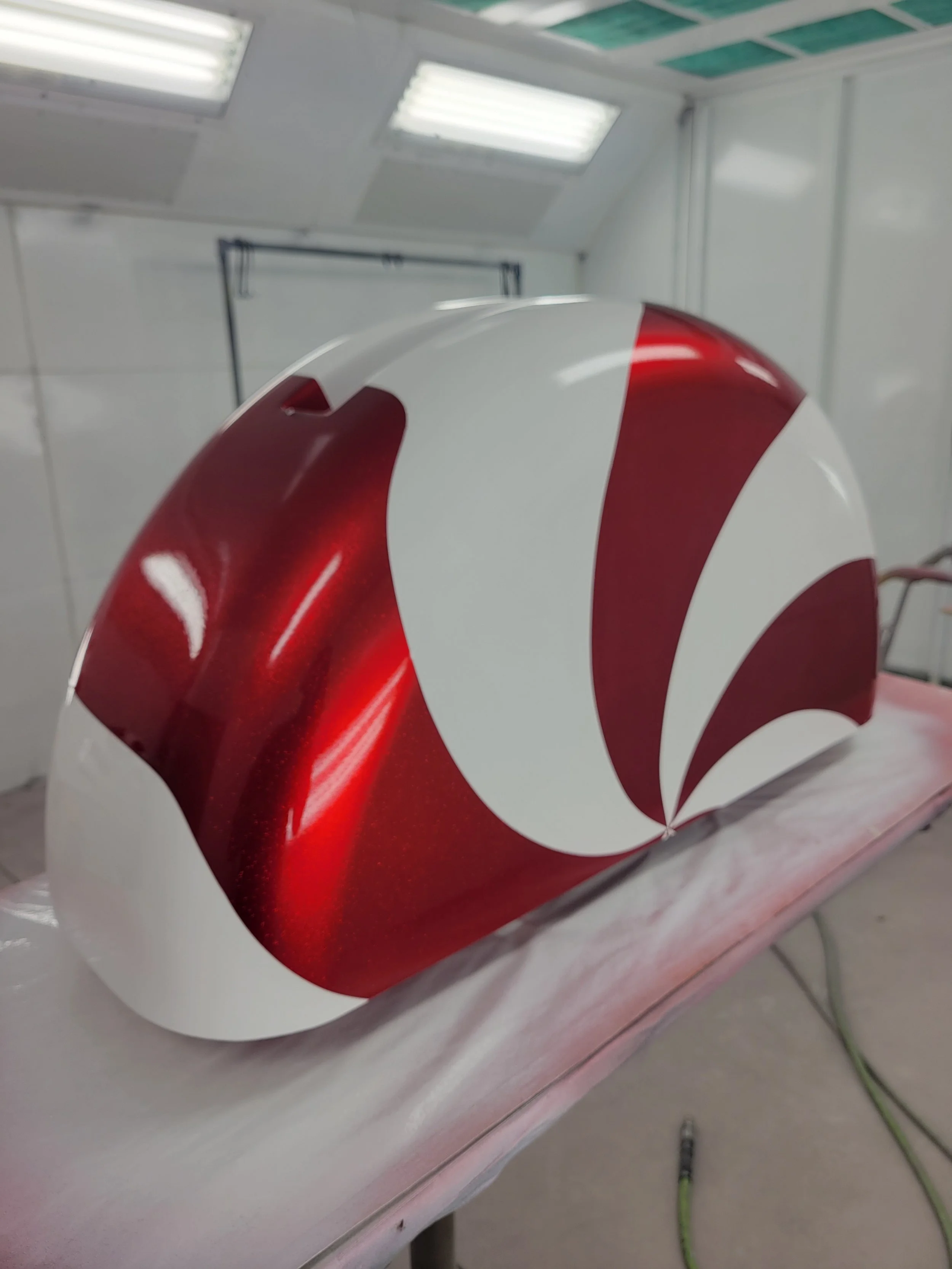 Close-up of a motorcycle fuel tank with a red and white swirl paint design, placed on a padded surface in a workshop or garage.