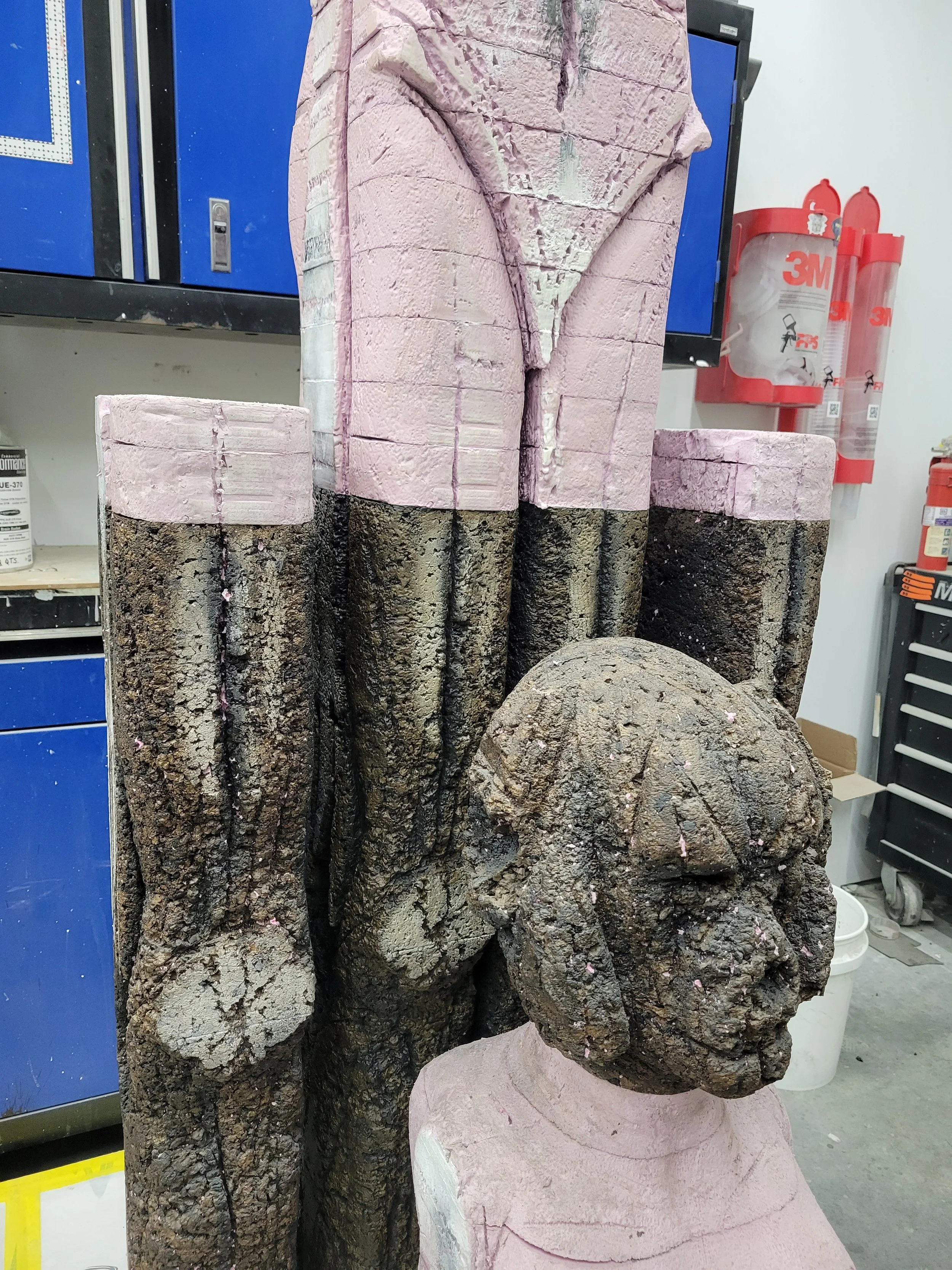 A large sculpture of a head and bust made of textured material resembling stone, with some parts painted pink, situated in a workshop environment.