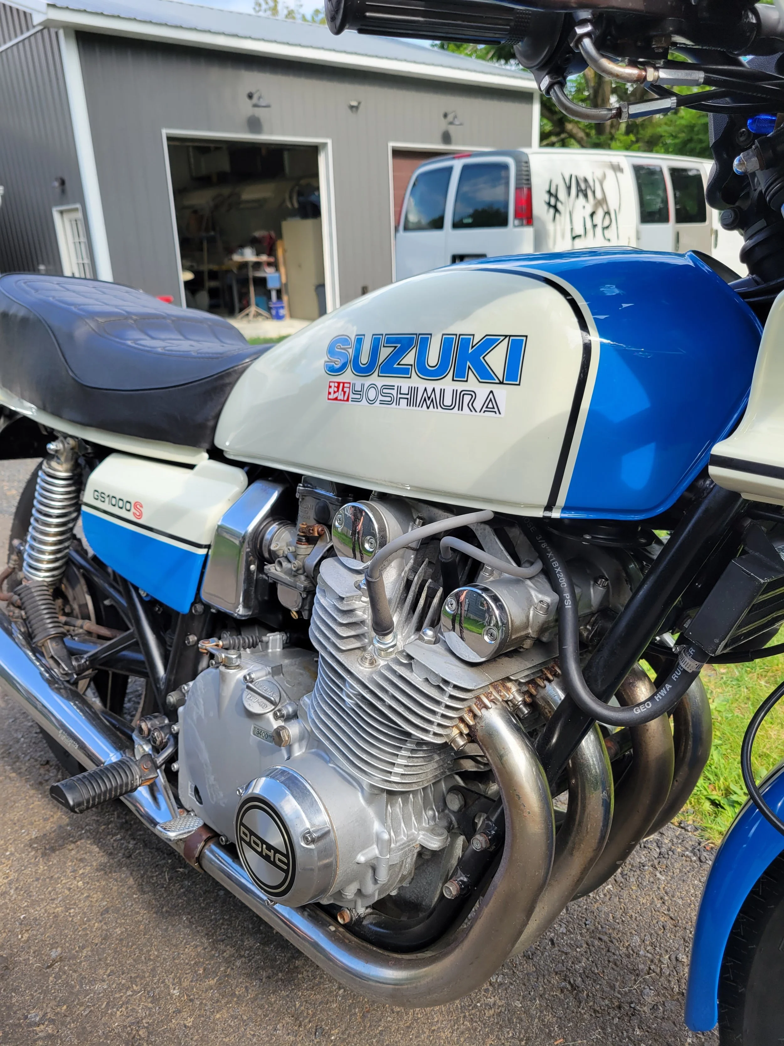 Close-up of a vintage Suzuki motorcycle with a white and blue fuel tank and engine, parked outdoors near a building and a van in the background.