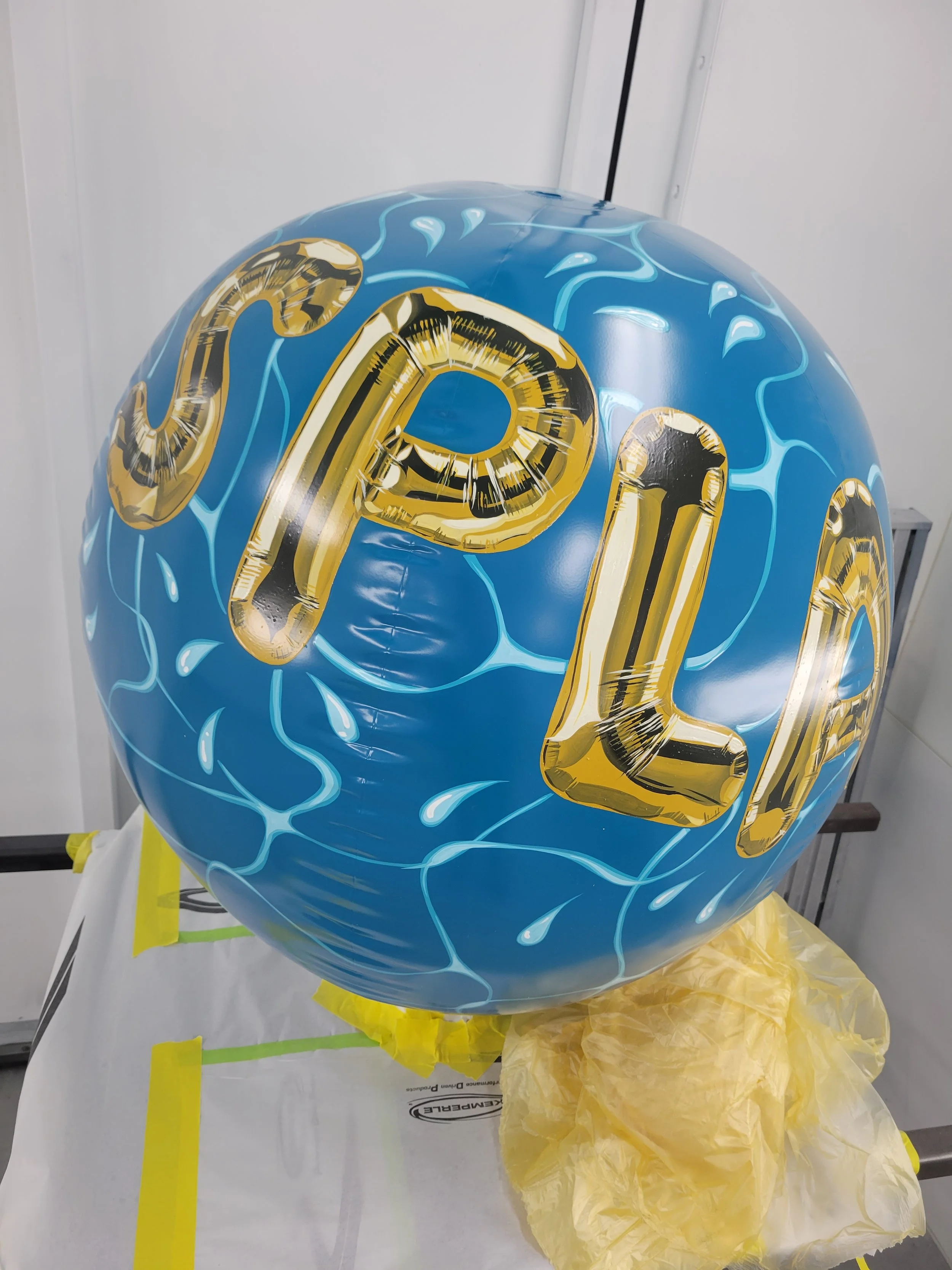 Blue water-themed inflatable ball with gold foil letters spelling 'SPLASH' on it, resting on yellow tissue paper.