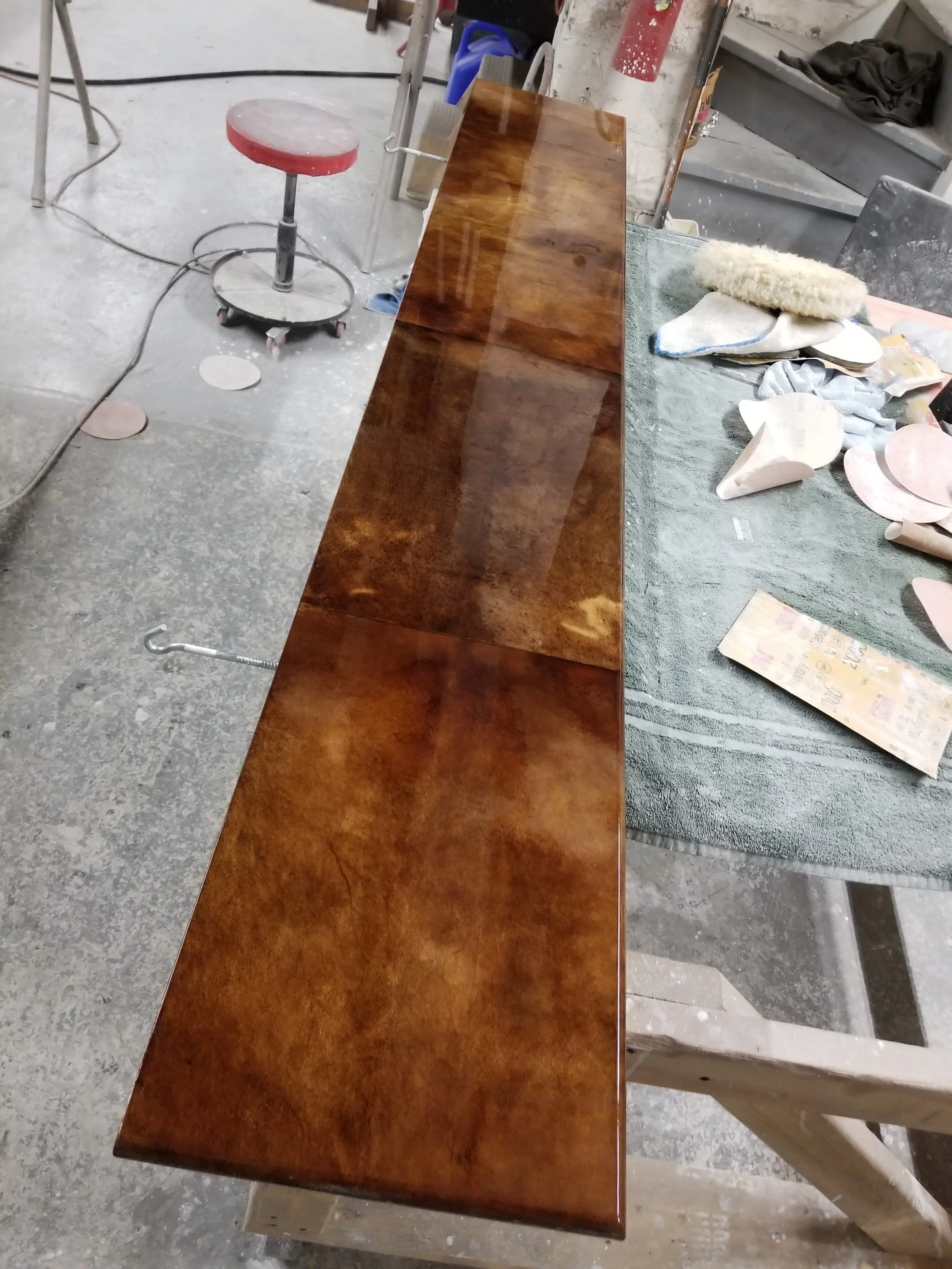 A long, polished wooden table in a workshop, with various tools, sanding pads, and cloths on the surface, and a red stool nearby.