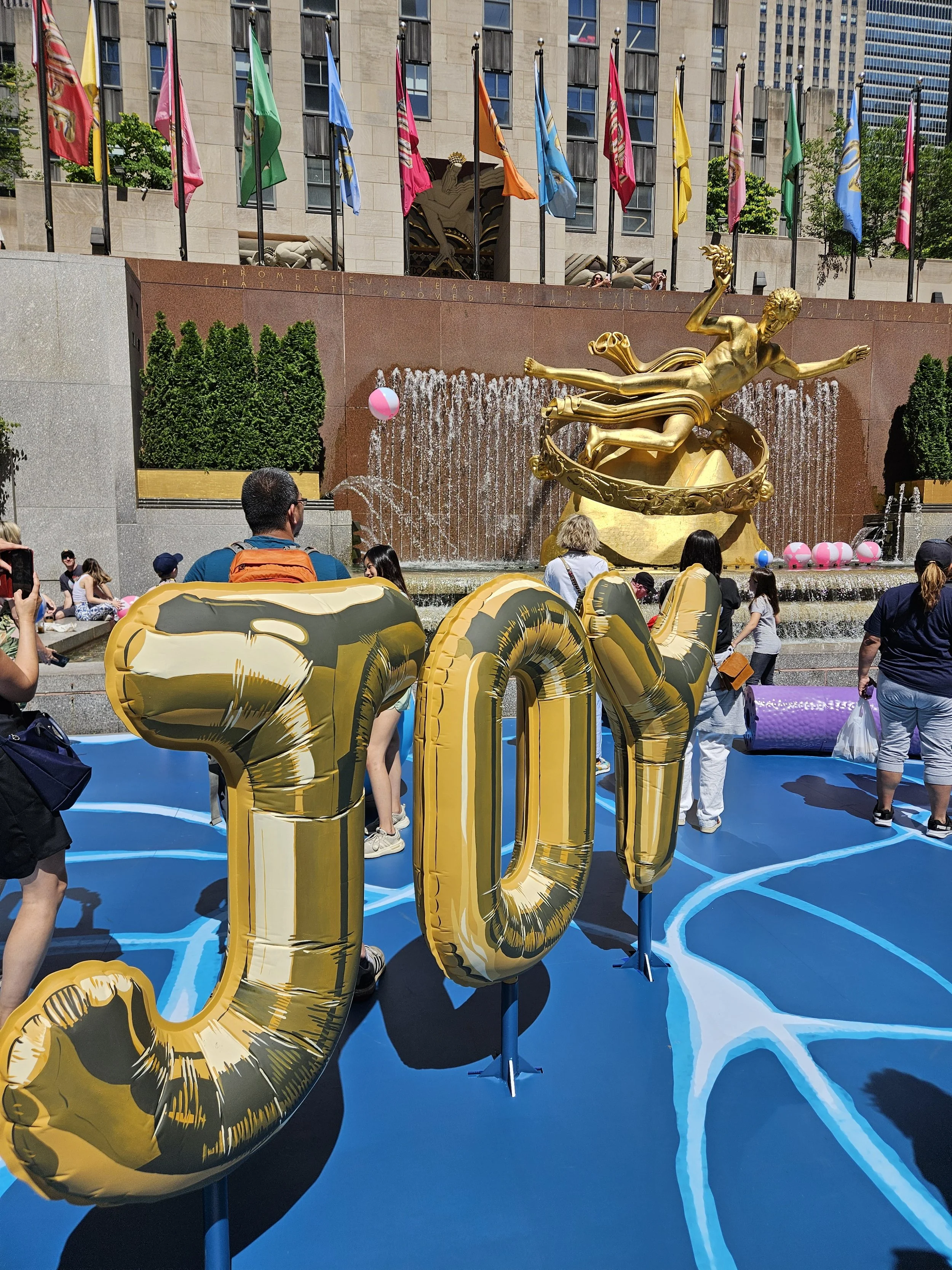 People gathering near a fountain with a large golden statue of a woman, surrounded by pink beach balls, and golden balloons spelling 'JOY' on a blue surface, with colorful flags in the background.