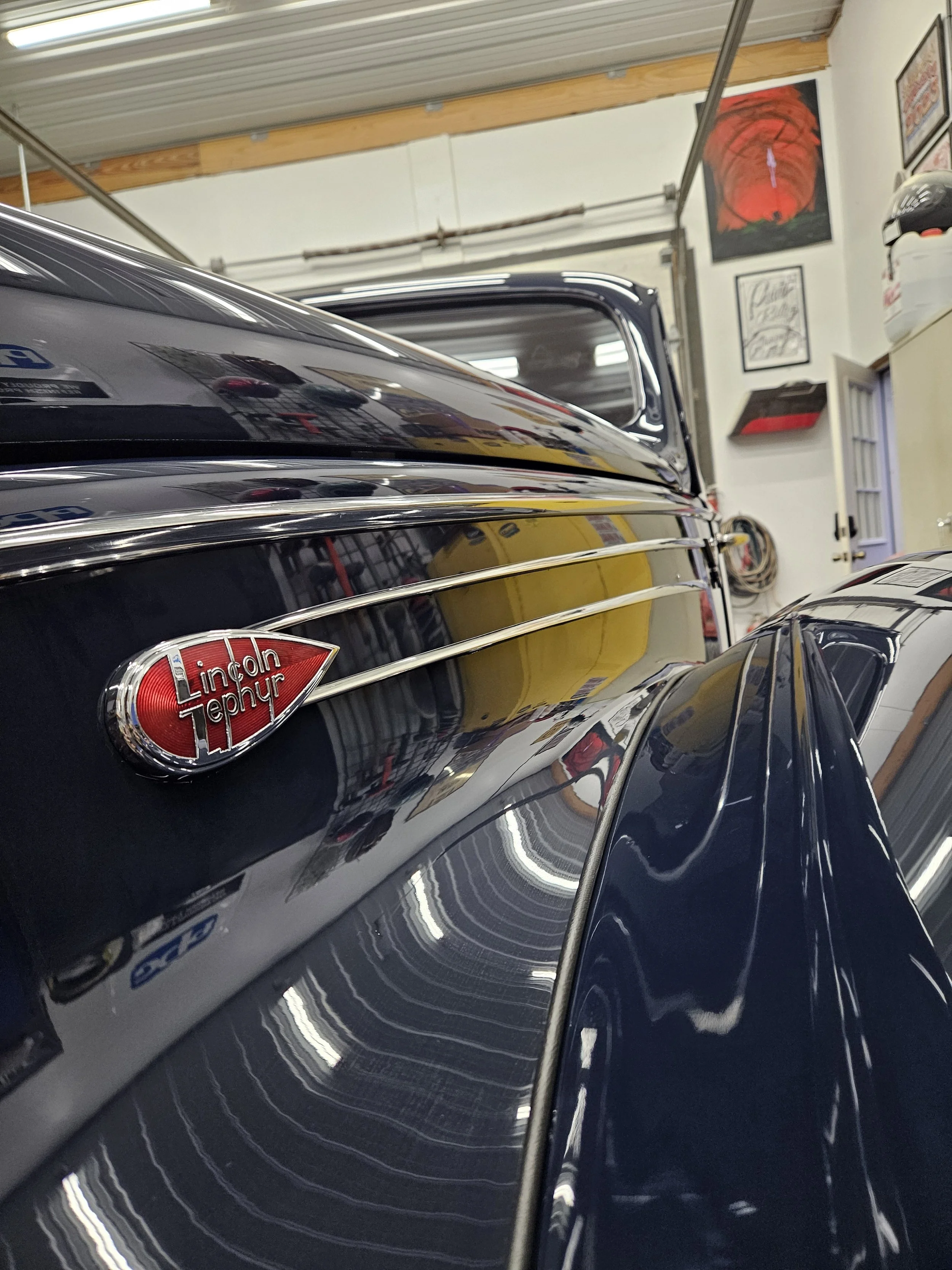 A close-up view of a vintage black Lincoln Zephyr car in a garage, showing the car's badge and reflections on its smooth surface.