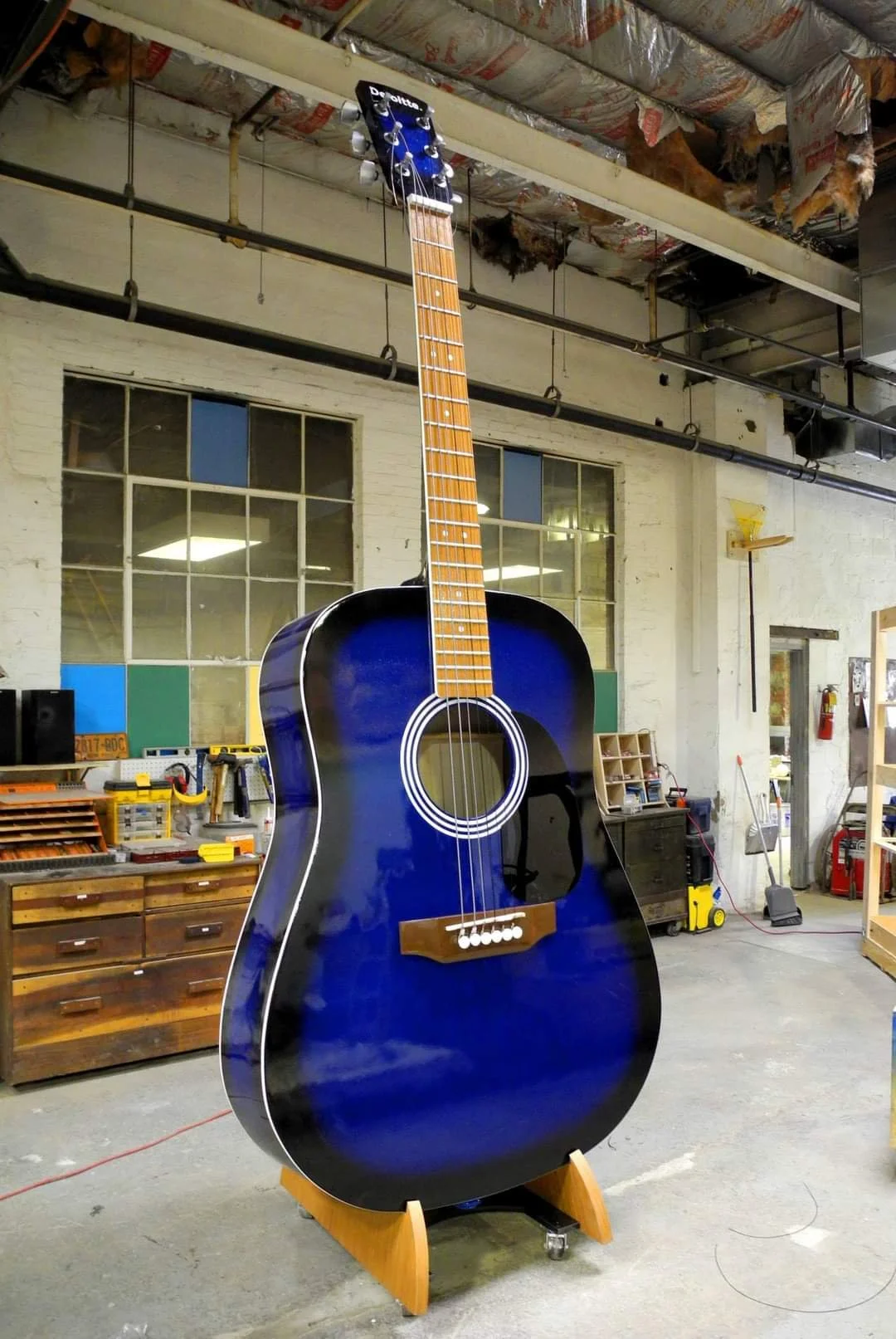 Blue acoustic guitar on a wooden stand in a workshop with tools, shelves, and a large window in the background.