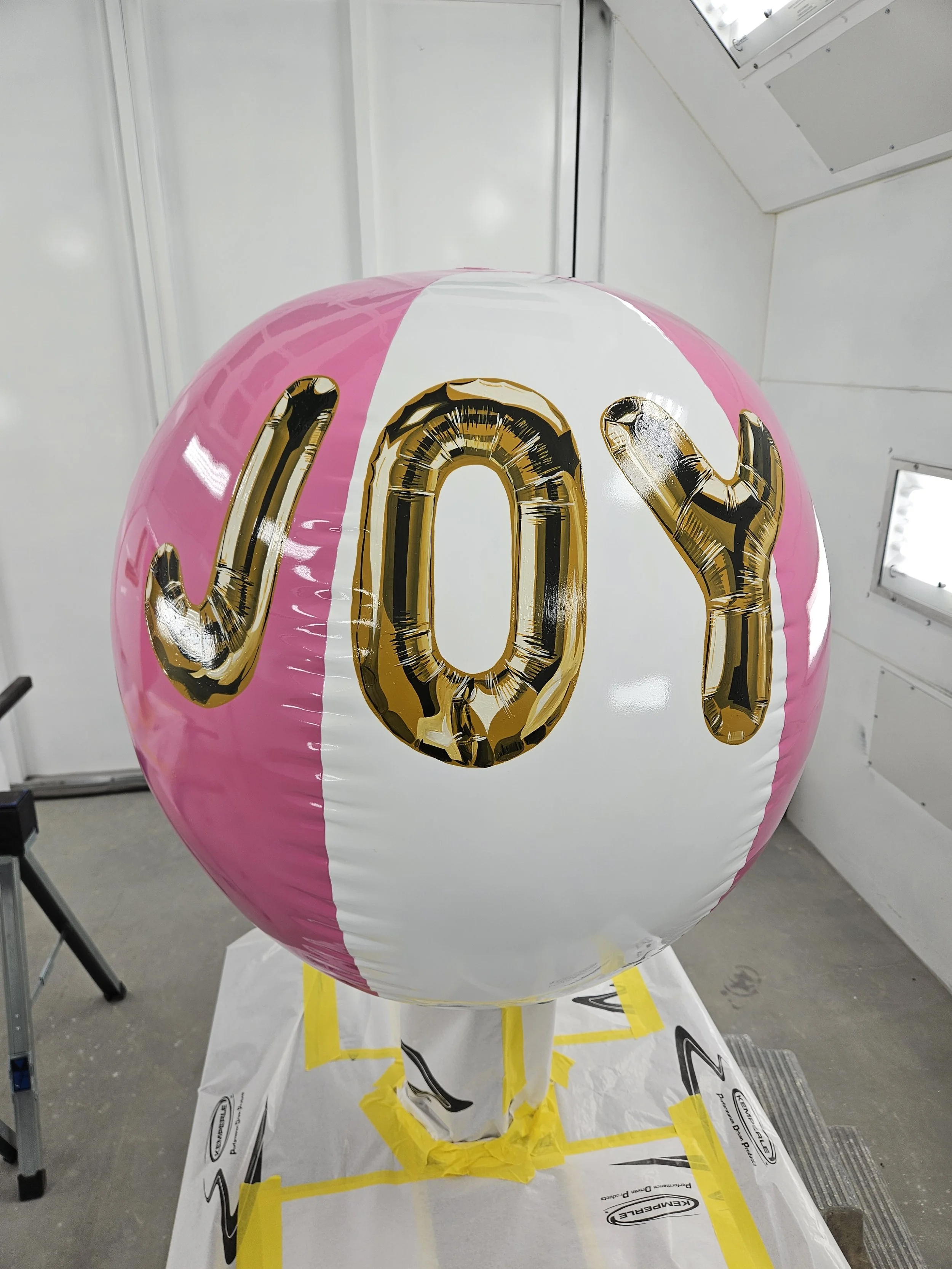 Pink and white beach ball with gold and black letter balloons spelling 'Joy' on it, placed on a stand covered with yellow tape inside a white room.