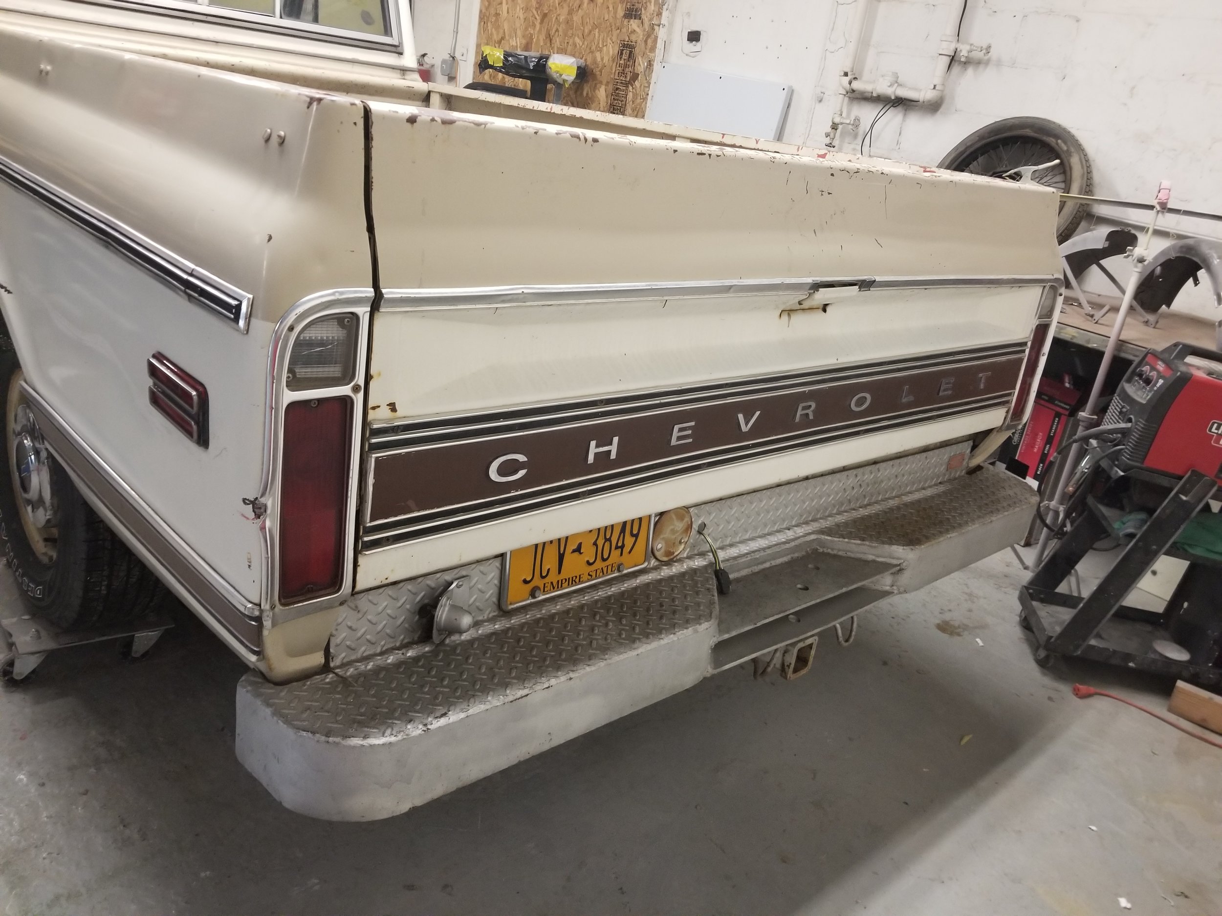 Rear view of an old Chevrolet pickup truck in a garage, showing the tailgate, rear lights, and bumper with a yellow license plate, surrounded by various tools and equipment.