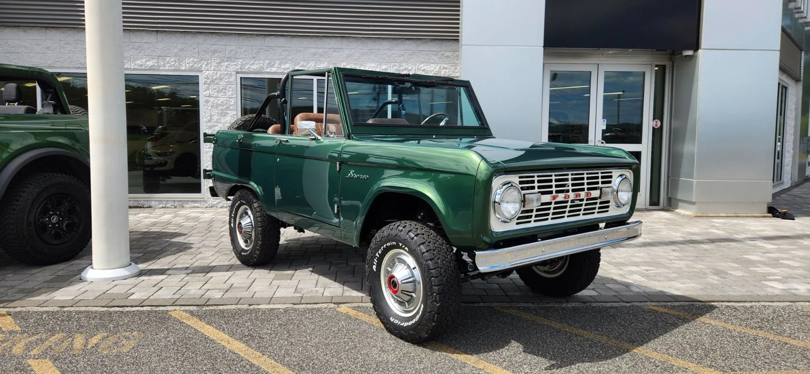A vintage green Ford Bronco SUV parked on a brick-paved lot outside a building with glass windows and a metal door.