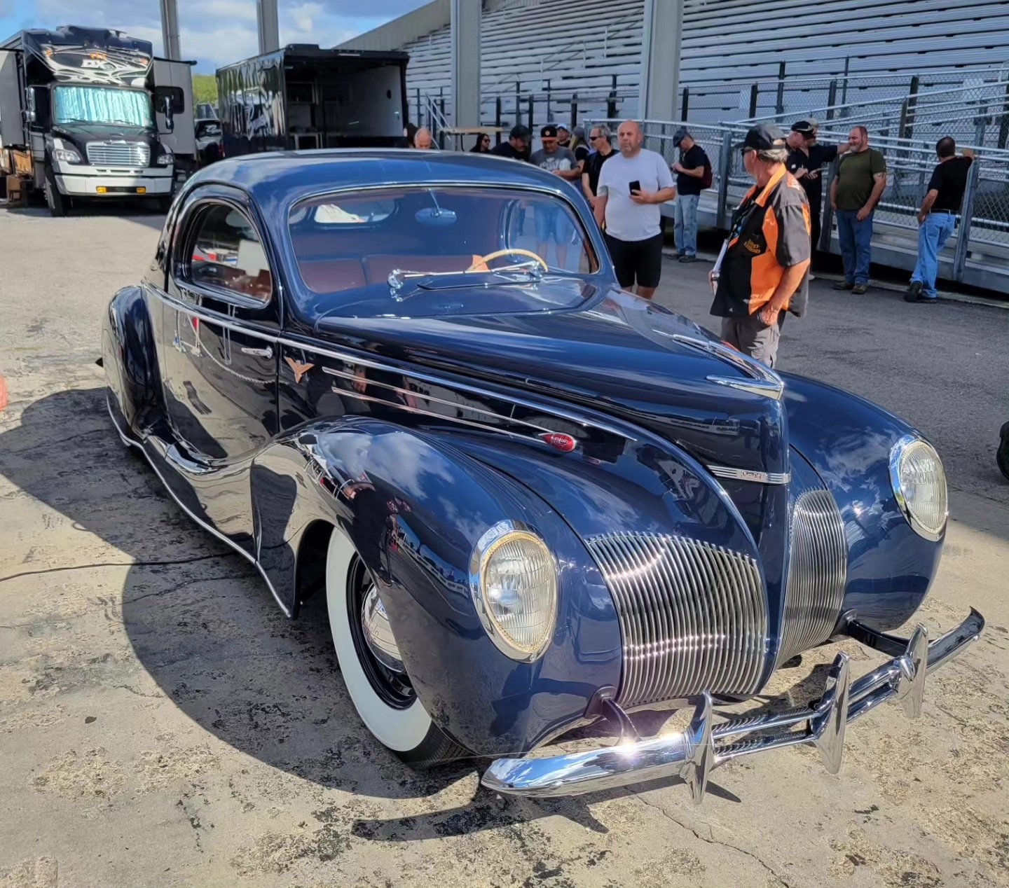A vintage black car with a shiny exterior and rounded design, parked at an outdoor event with people in the background.