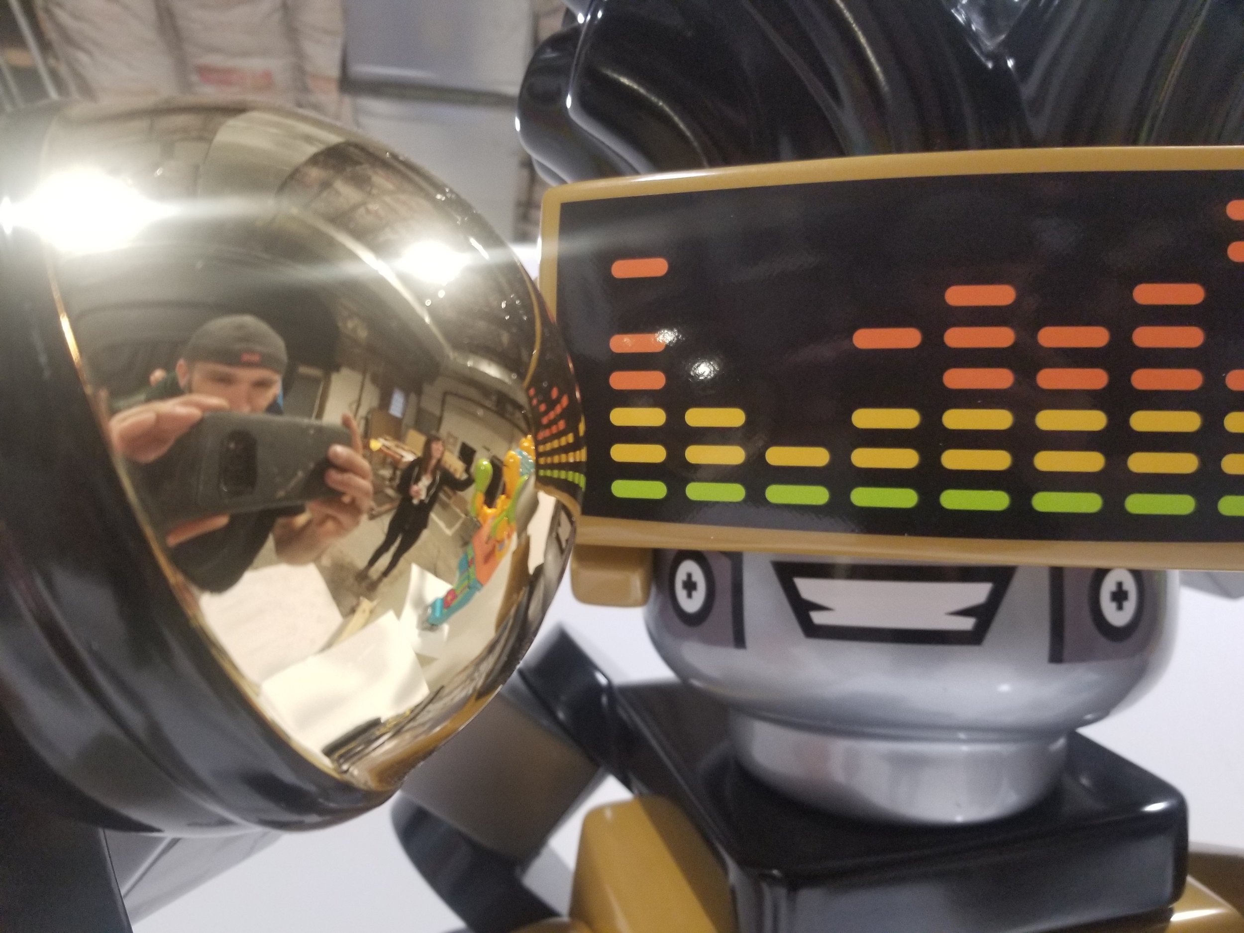 Close-up of a robot with a display showing colorful horizontal bars. The robot's head has a shiny, reflective surface, showing a person taking a photo with a smartphone in the reflection. The background shows an indoor setting with a person and vario