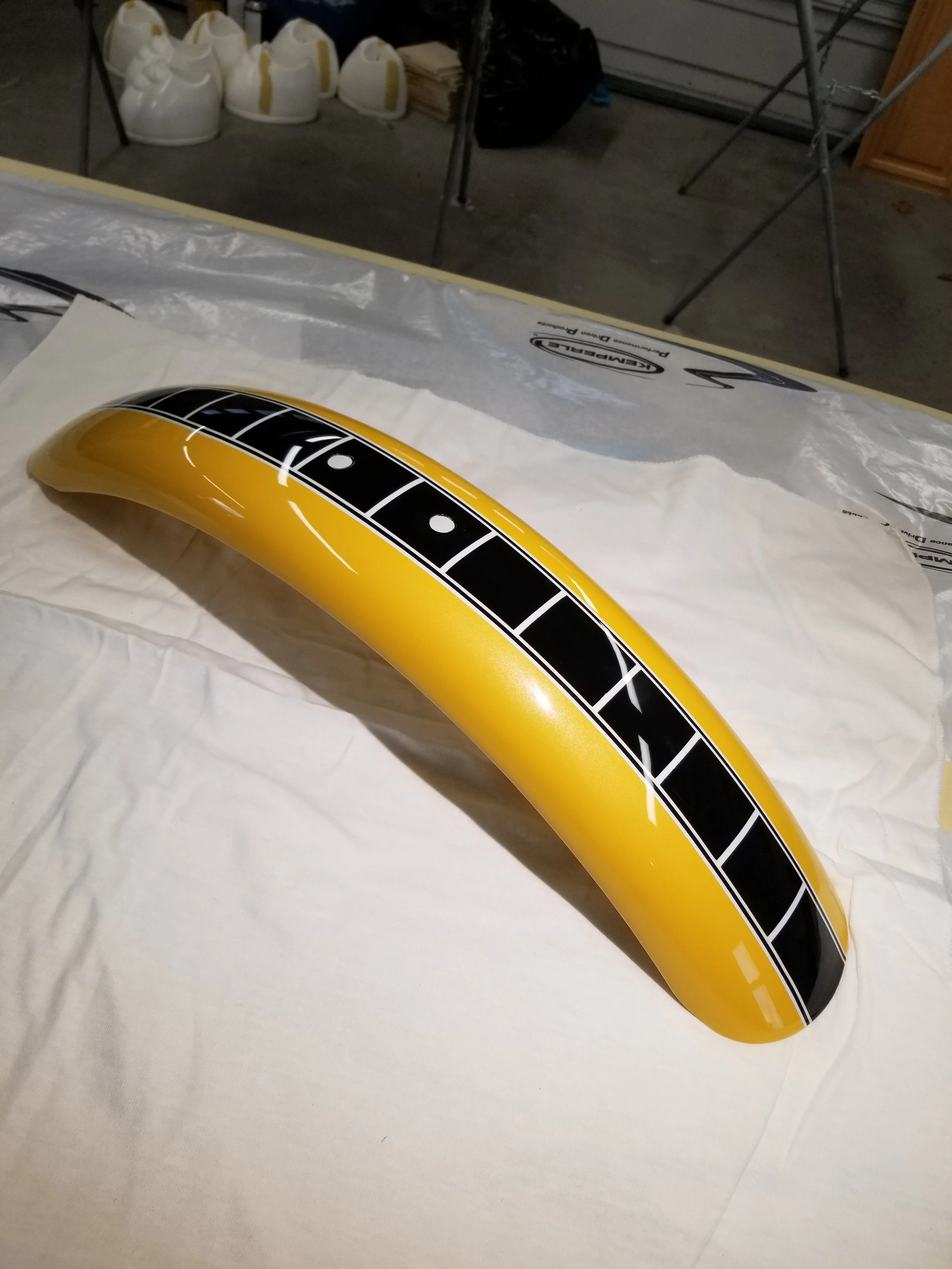 A custom painted yellow motorcyle part with black and white designs resting on a white cloth, with a garage or workshop background.