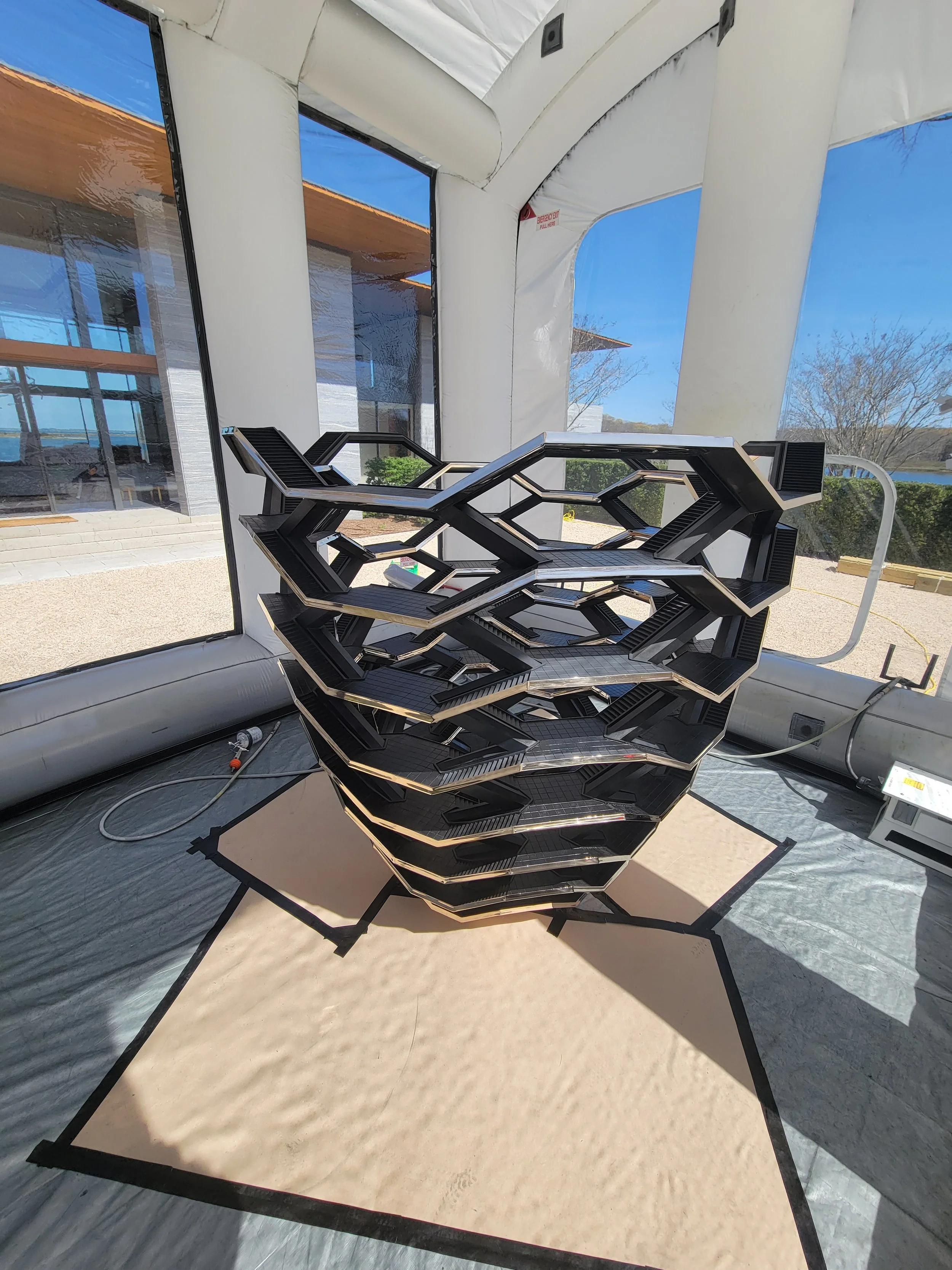 Stacked black and silver architectural model or structure inside a glass enclosure, with large windows showing a clear blue sky and outdoor landscape.