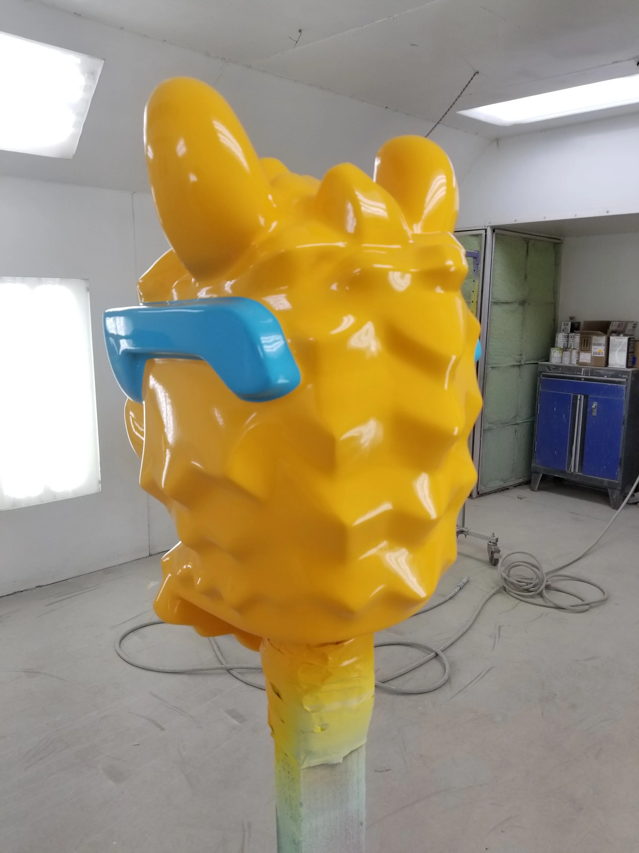 A large, yellow, LEGO camel head with blue sunglasses on a stand in a workshop.