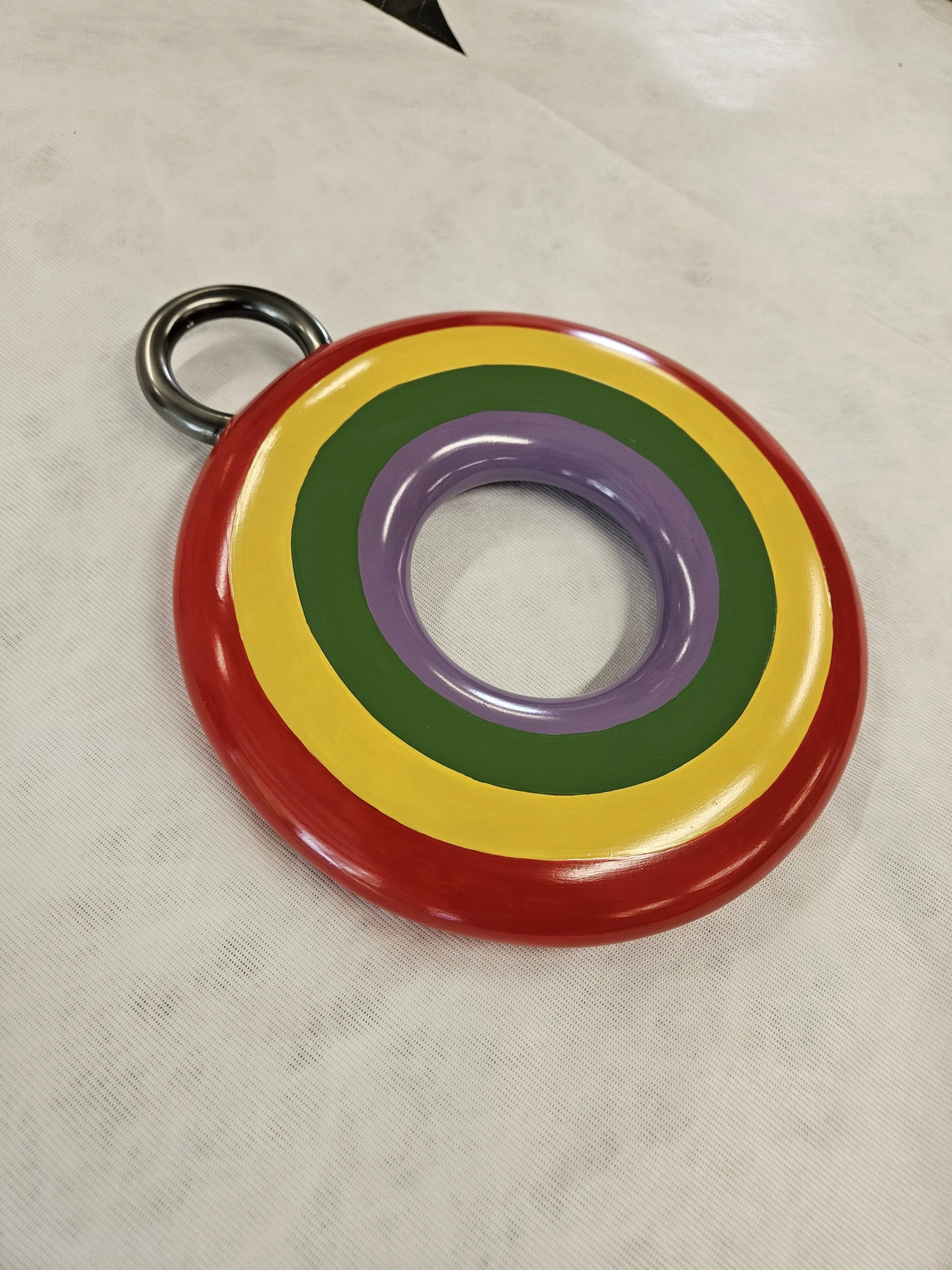 Colorful rubber ring with red, yellow, green, and purple concentric circles, and a metal loop attached to the edge, resting on a white surface.