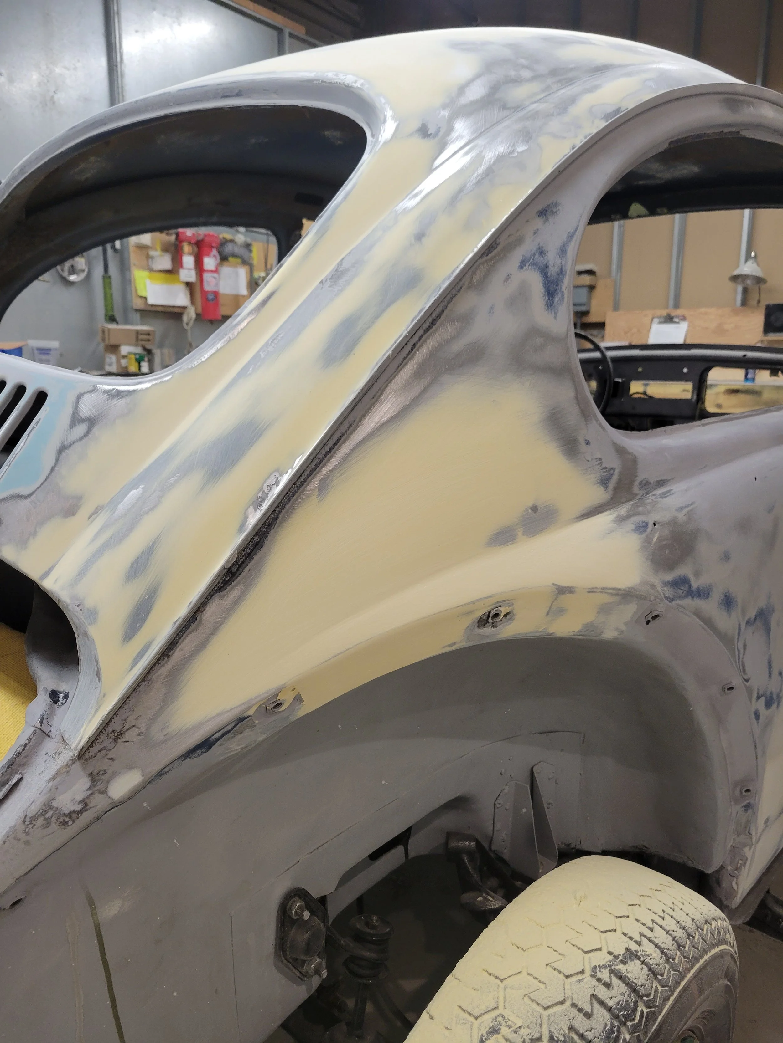 Partial view of a vintage car in the process of restoration, with the body prepped and sanded, showing areas of primer and body filler.